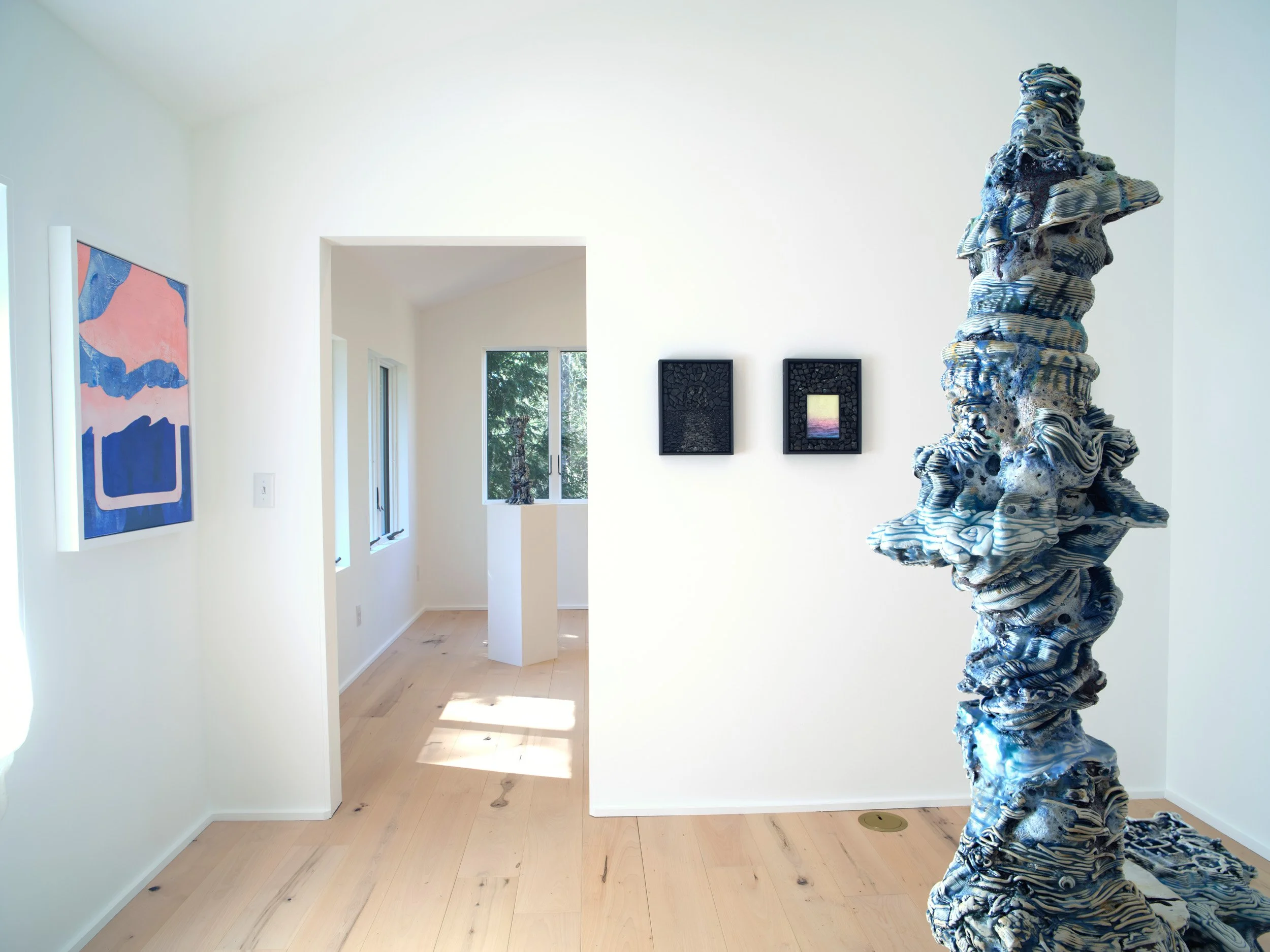 Interior of a bright art gallery with abstract paintings on white walls, a sculpture with swirling blue, gray, and black patterns in the foreground, and a window showing trees outside.