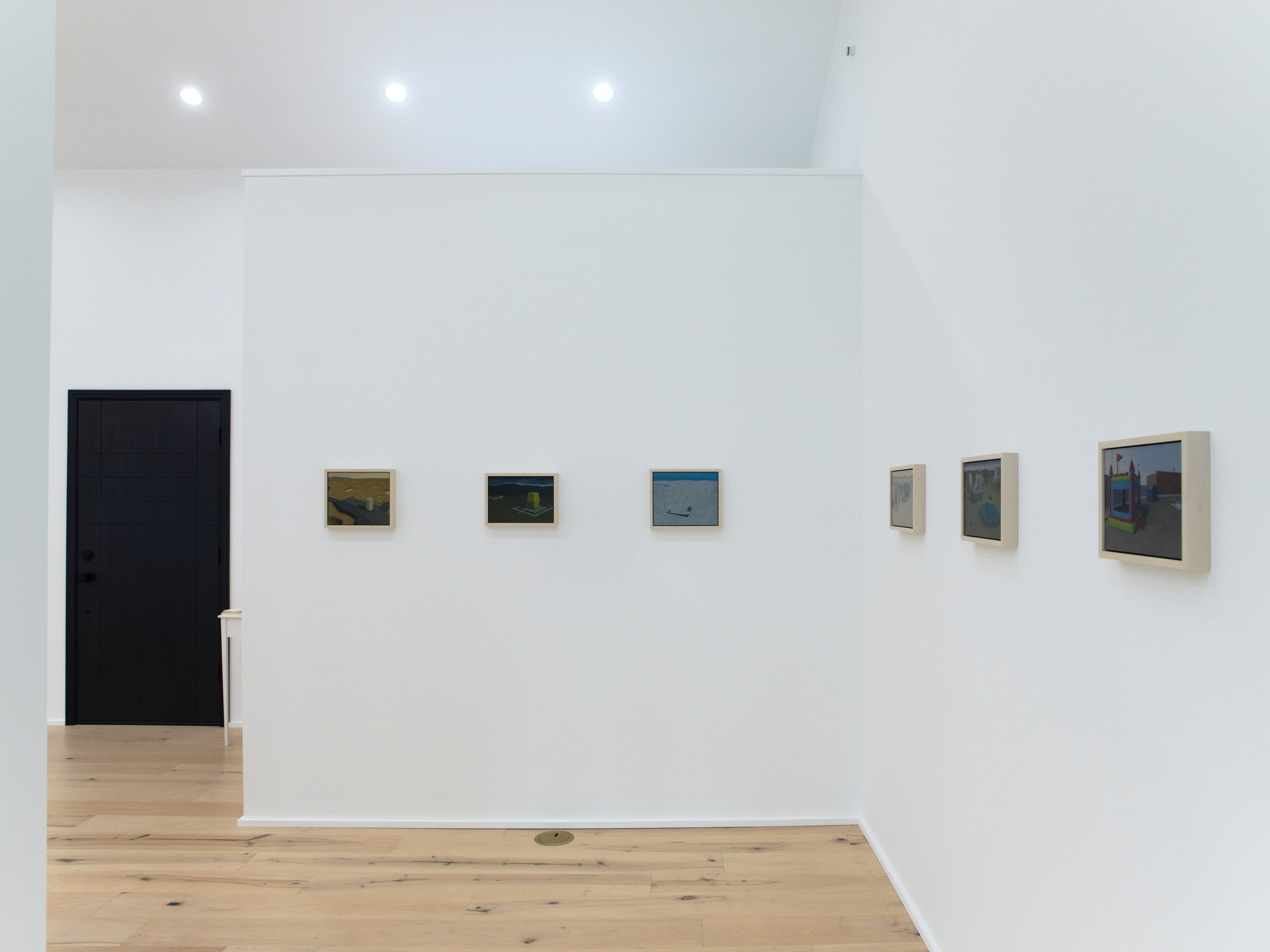 An art gallery with white walls featuring framed pictures hung in a row. A black door is on the left and wooden floors are visible.