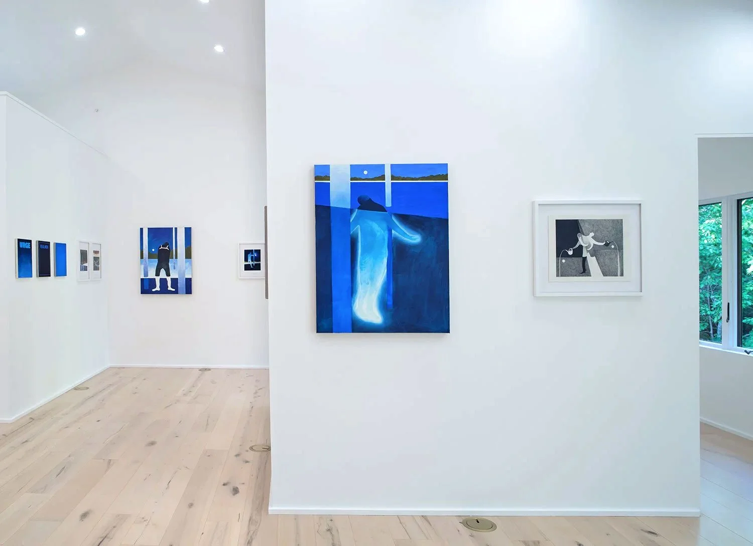 Gallery wall displaying various modern art paintings in white frames, with a focus on a large blue abstract piece and smaller artworks on adjacent walls, natural light from a window illuminating the space.