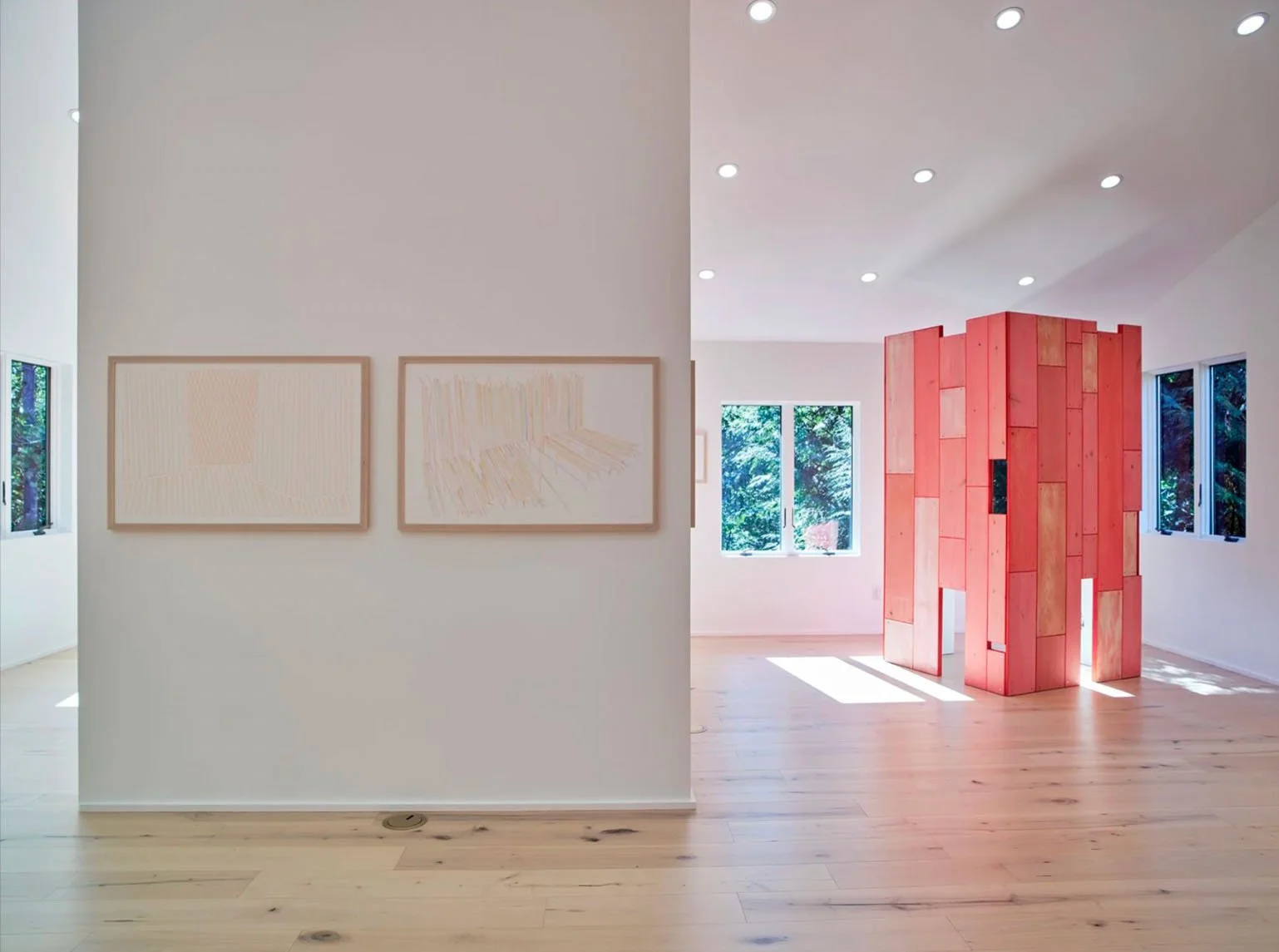 An art gallery features two abstract paintings on a white wall and a wooden partition with a geometric design in the background, sunlight coming through windows.