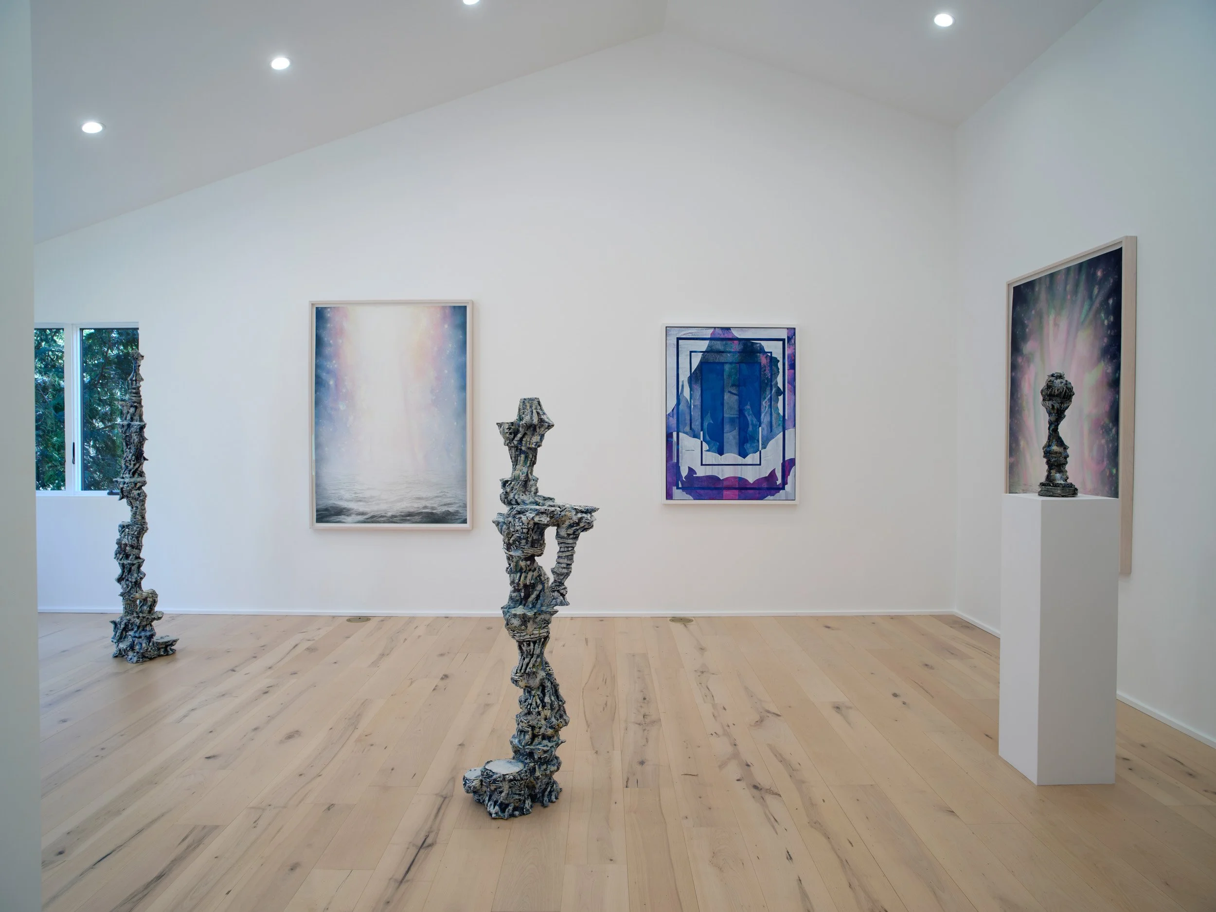 An art gallery with a white wall, framed artworks, and sculptural pieces on pedestals in the foreground. Wooden flooring and a window showing outdoor greenery.