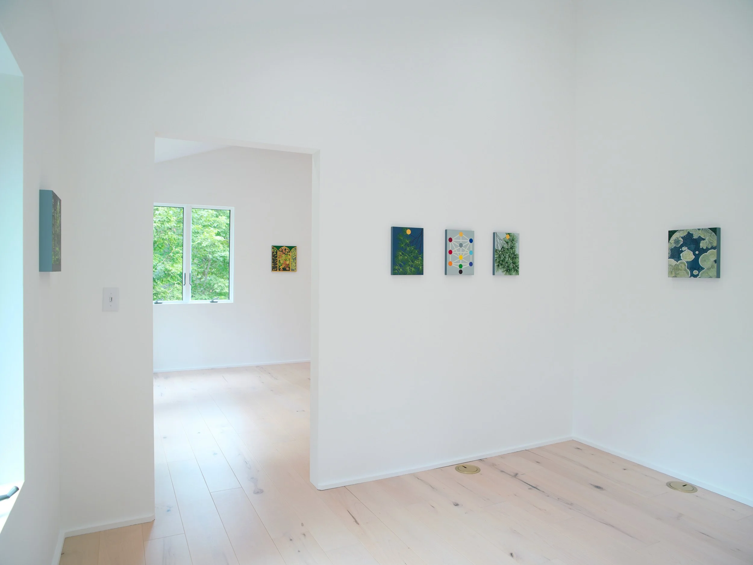 Interior view of a bright, minimalistic room with white walls, light wooden flooring, and a window showing green foliage outside. Several framed artworks are hung on the wall.