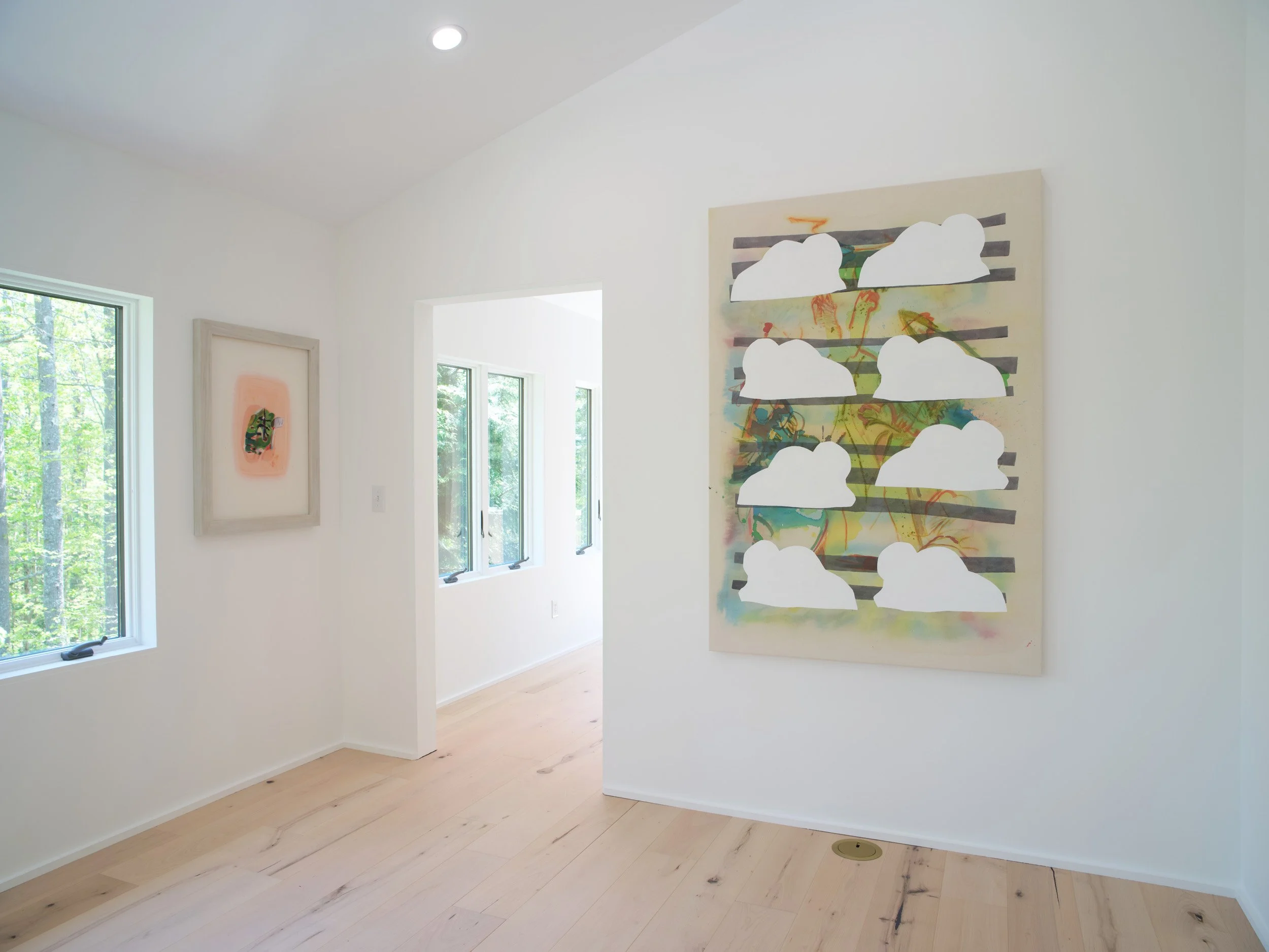 Interior of a bright art gallery with colorful abstract paintings on white walls and large windows showing green trees outside.