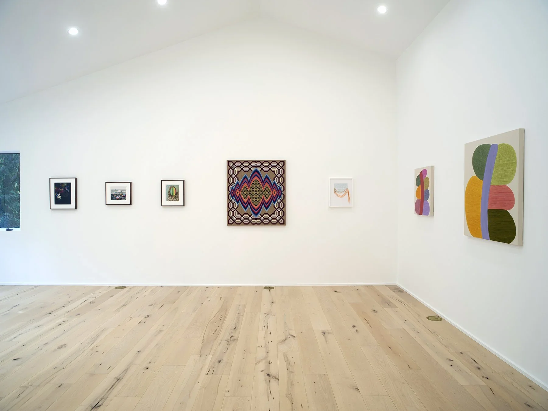 Art gallery with white walls displaying colorful abstract and patterned artworks, wood flooring, and ceiling lights.