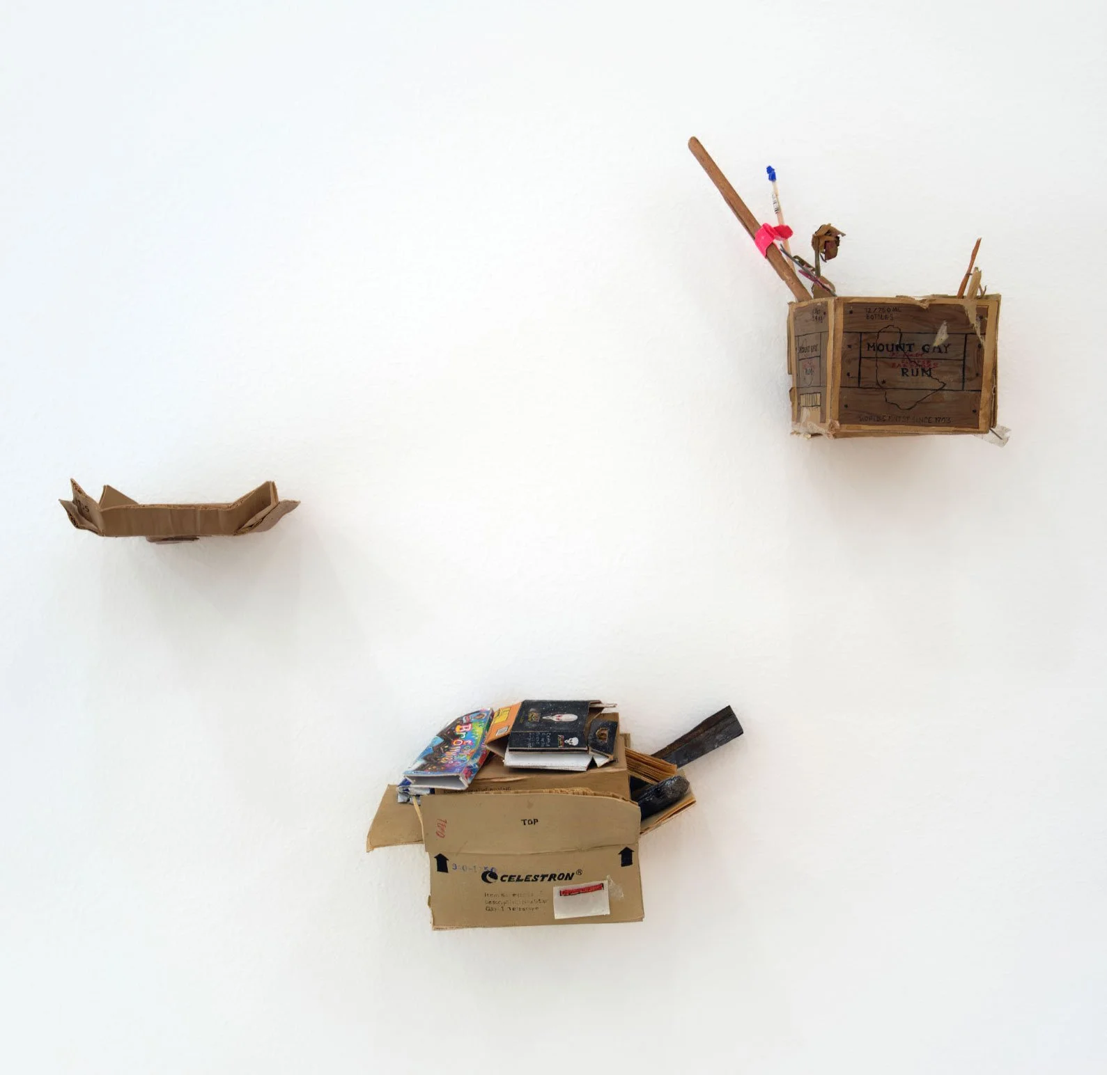 Mixed objects on a white wall: a small empty cardboard boat, a pile of books in a cardboard box, a wall-mounted wooden crate with writing, and a wall-mounted wooden object.