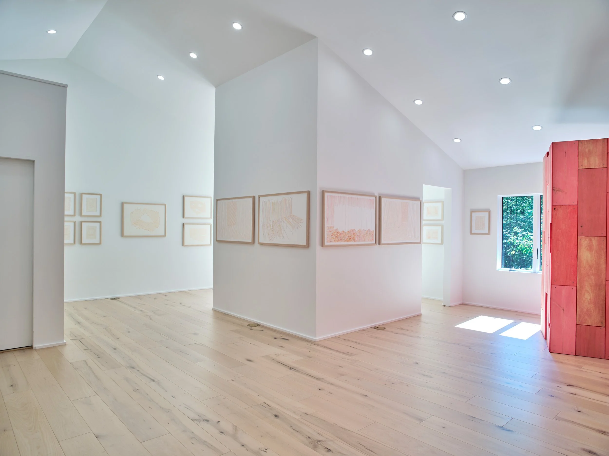 A contemporary art gallery with white walls, light wooden floors, and framed artwork on the walls. Natural light streams in through a window, and there is a red partition on the right side.