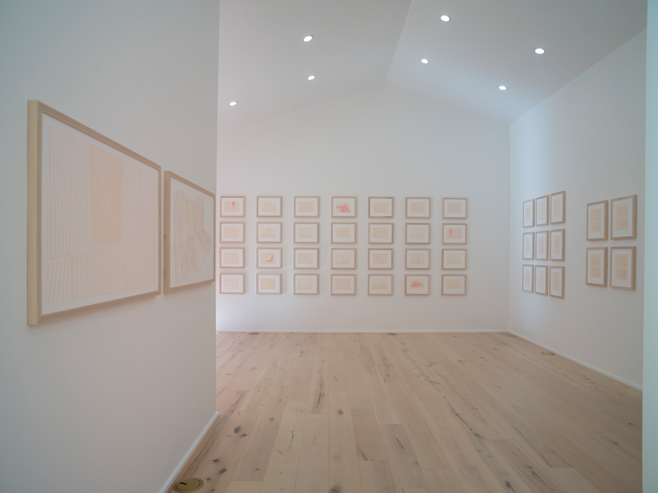 An art gallery with framed artwork displayed on white walls and ceiling spotlights, wooden floor, and a doorway leading to another room.