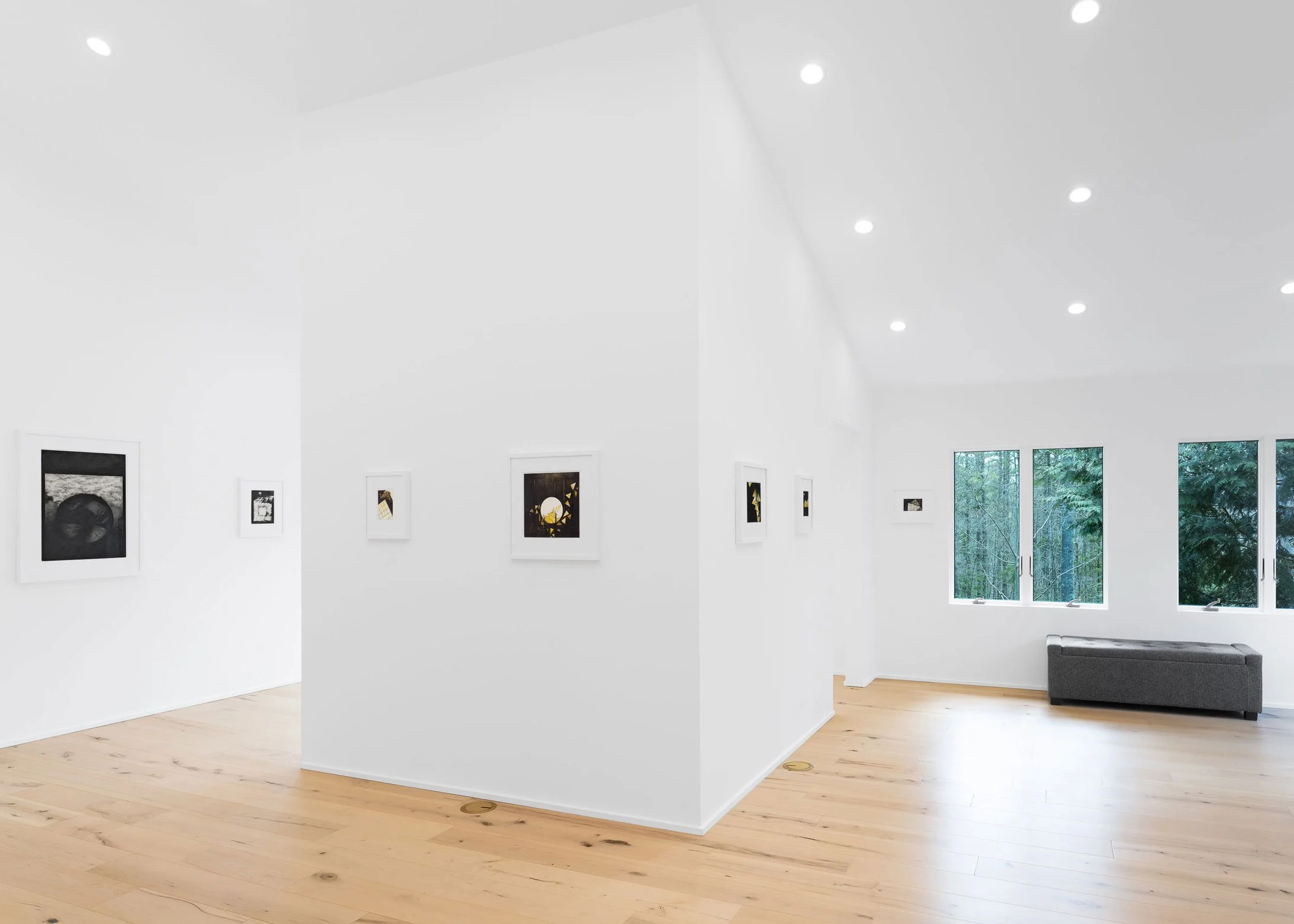A modern art gallery with white walls and light wood flooring, featuring framed black and white photographs, sunny windows with trees outside, and a gray sofa in the corner.