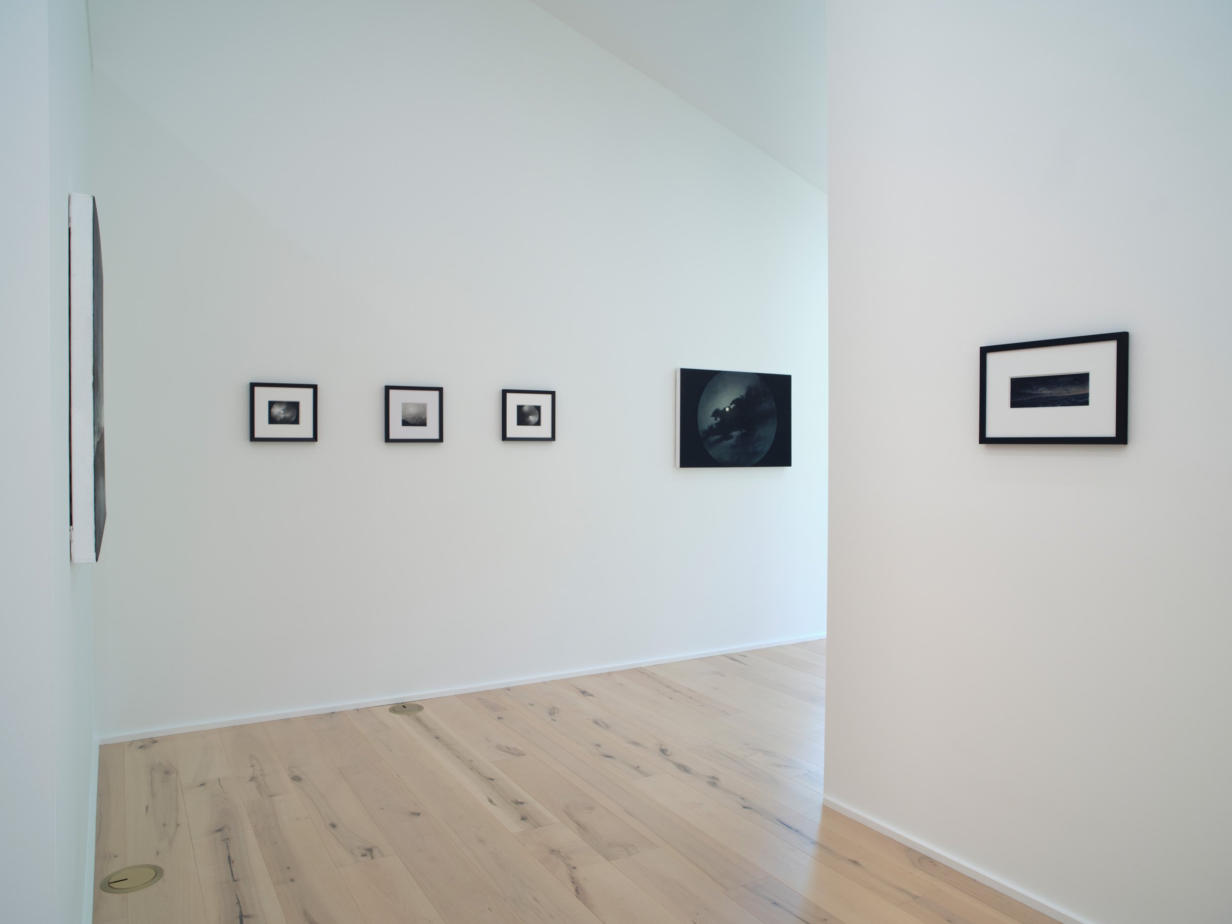 An art gallery with white walls displaying black and white framed photographs, light wood flooring, and minimal decor.