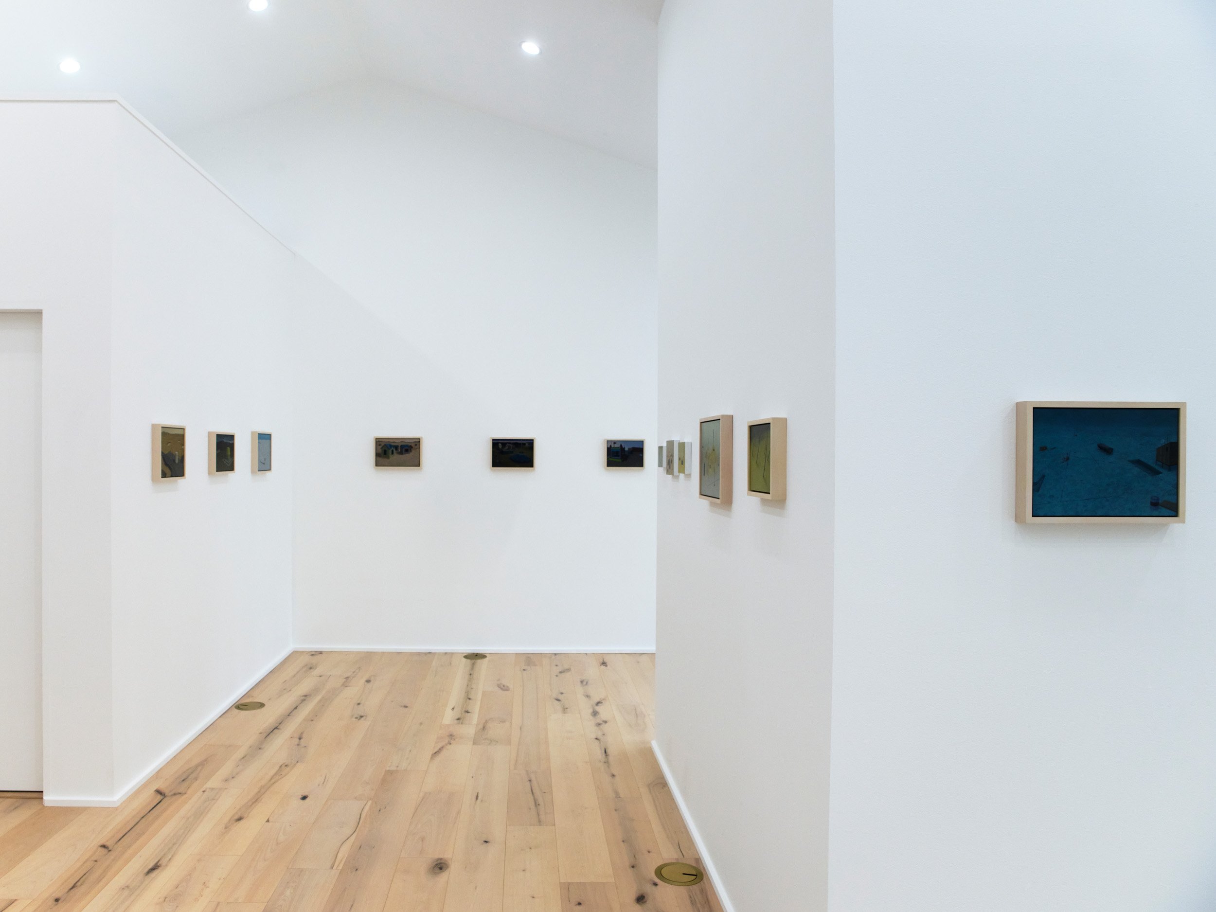 A modern art gallery with white walls displaying framed artworks. The gallery has wooden flooring, and there are ceiling lights illuminating the space.