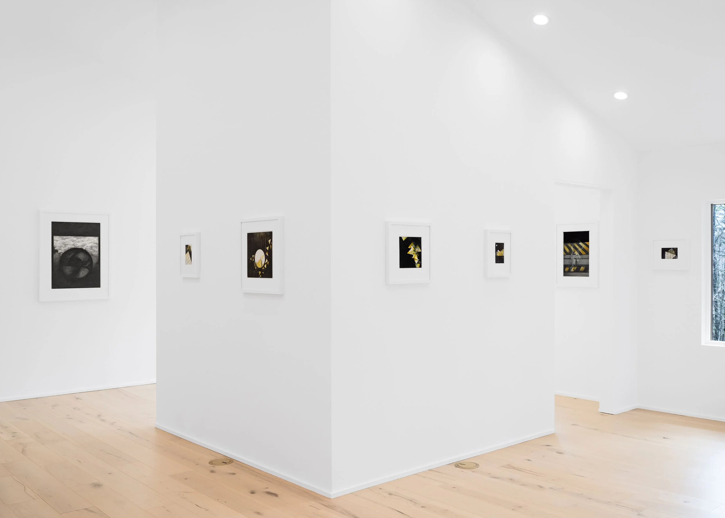 Interior view of an art gallery with white walls displaying framed artworks, wooden flooring, and ceiling lights. A window shows a glimpse of the outside.