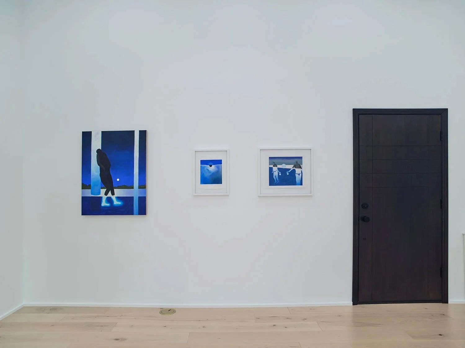 Gallery wall with three framed blue-toned artworks and a black door on a white wall and light wood floor.