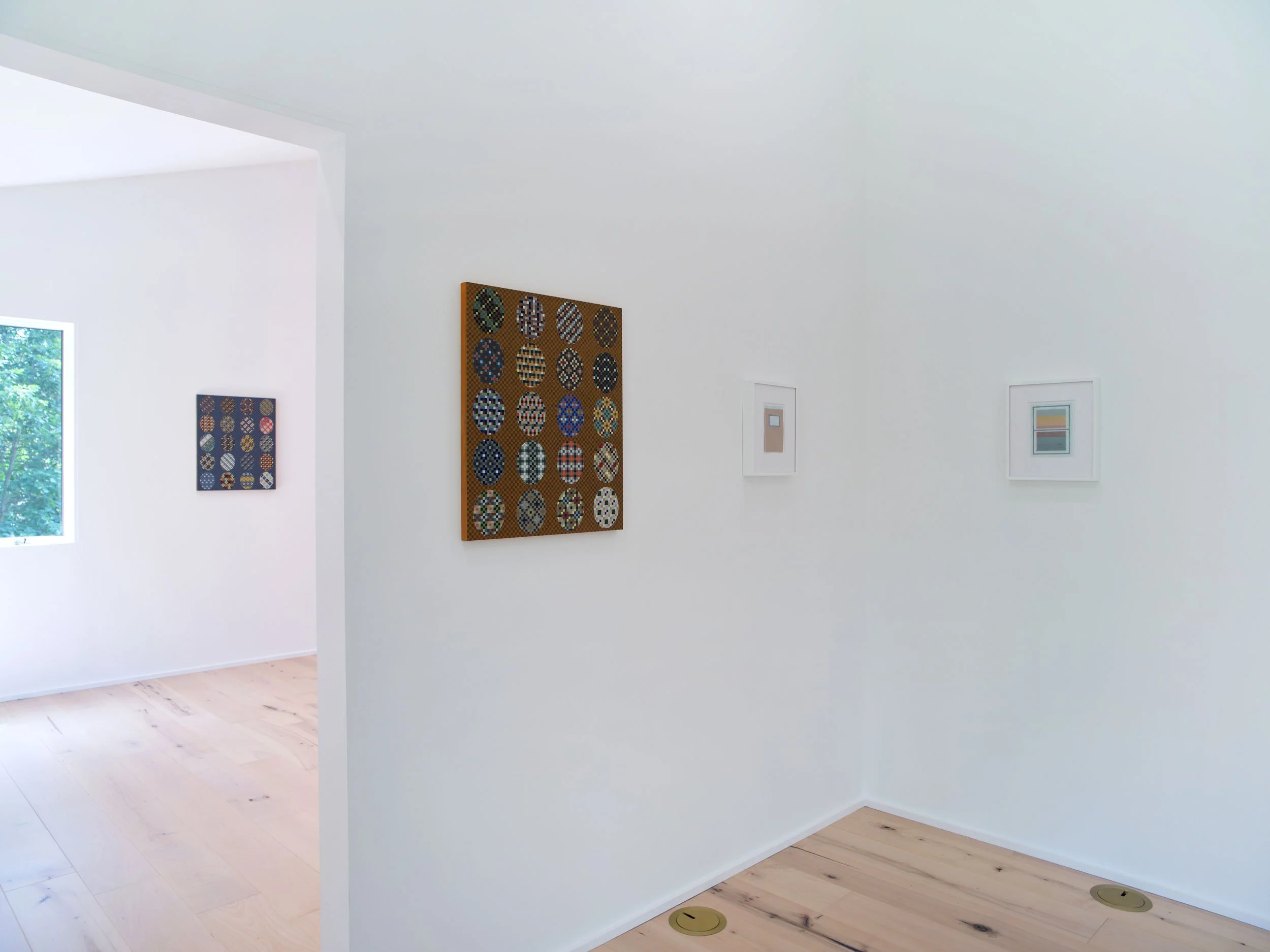 View of an art gallery with white walls displaying framed artwork and a large colorful piece made of circles. Natural light coming from a nearby window creates a bright atmosphere.