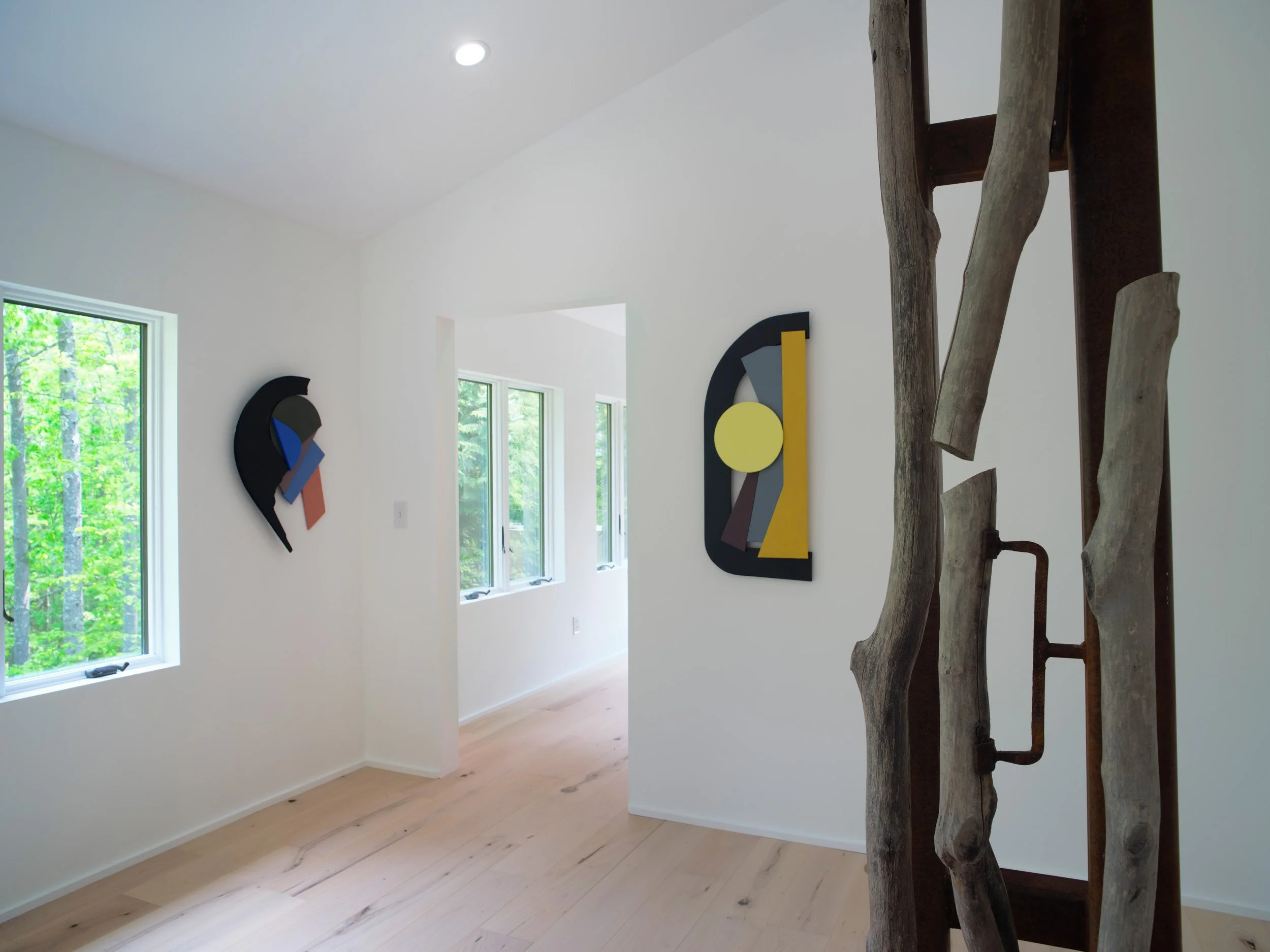 A bright, modern interior with white walls, wooden floors, and large windows showing green trees outside; wall art features abstract shapes and colors, and a natural wood sculpture or driftwood decor is visible in the foreground.