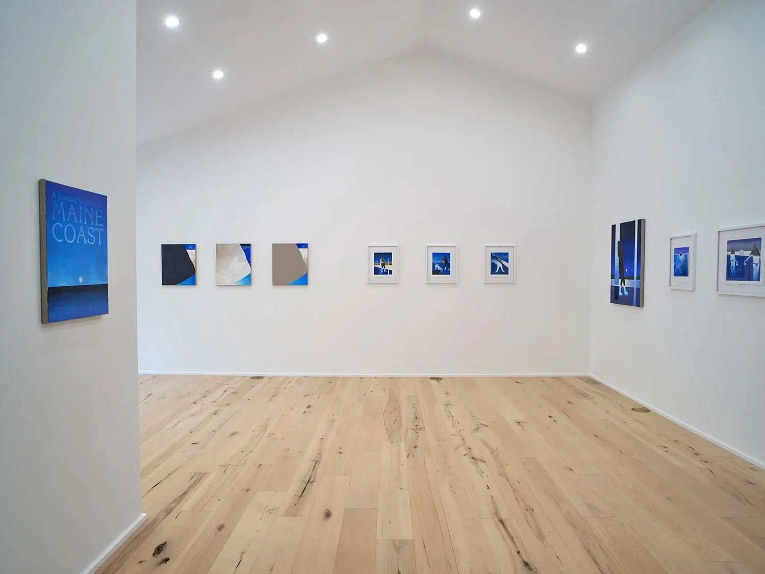 Art gallery with white walls and light wooden floor, displaying framed and canvas blue-themed artworks, including Maine Coast poster and various abstract and figurative images.
