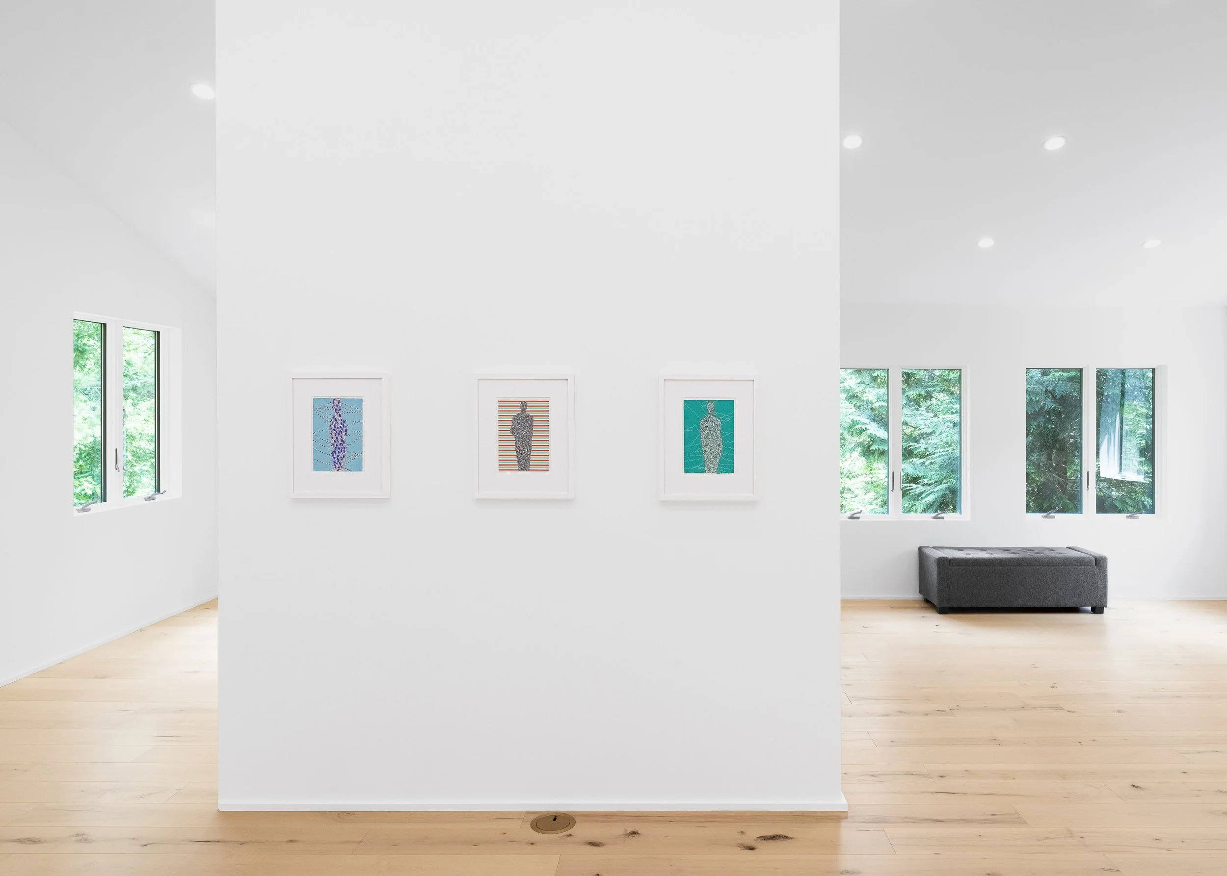 Contemporary art gallery with white walls and light wood flooring, featuring three framed artworks of abstract human figures hanging on a central white partition, with large windows showing greenery outside and a gray sofa on the right side.