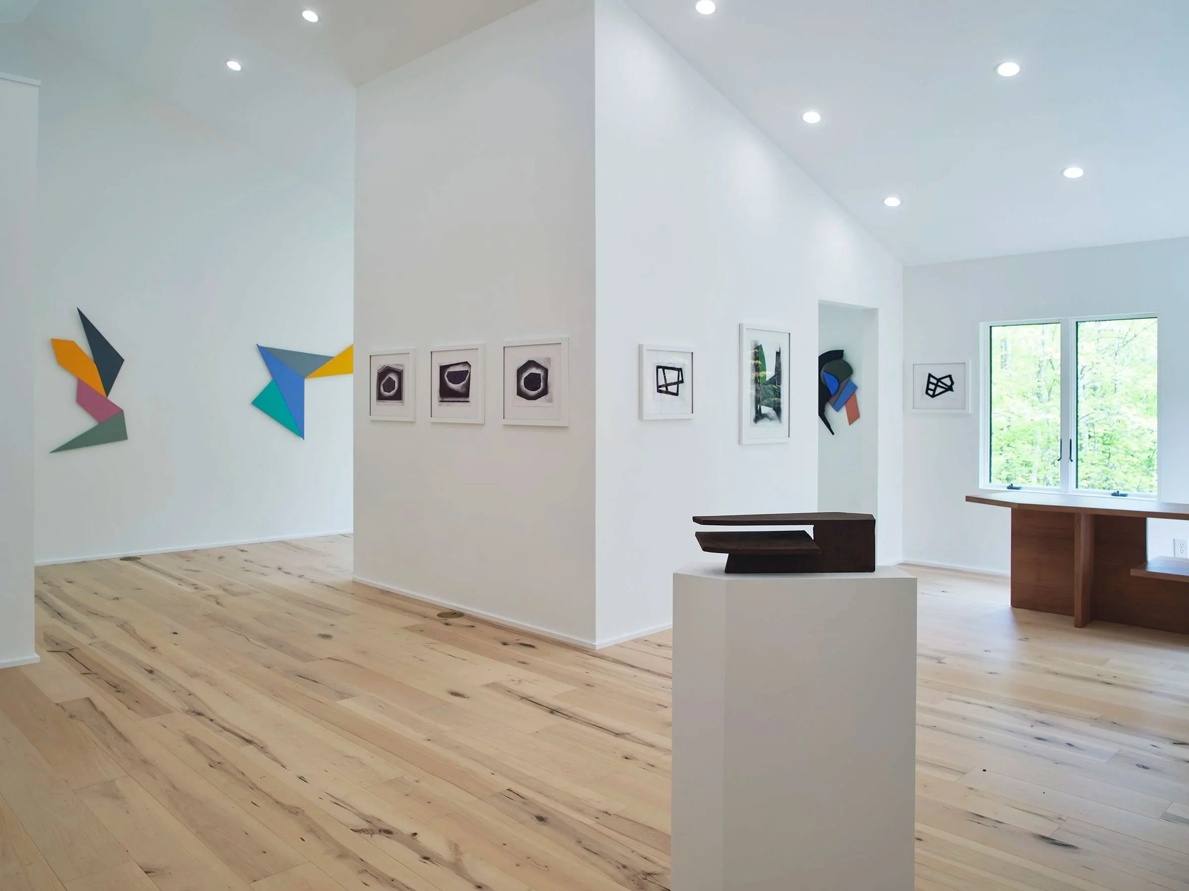 Interior view of an art gallery with white walls, wooden flooring, and various framed and abstract artworks displayed. Natural light through a window enhances the bright space.