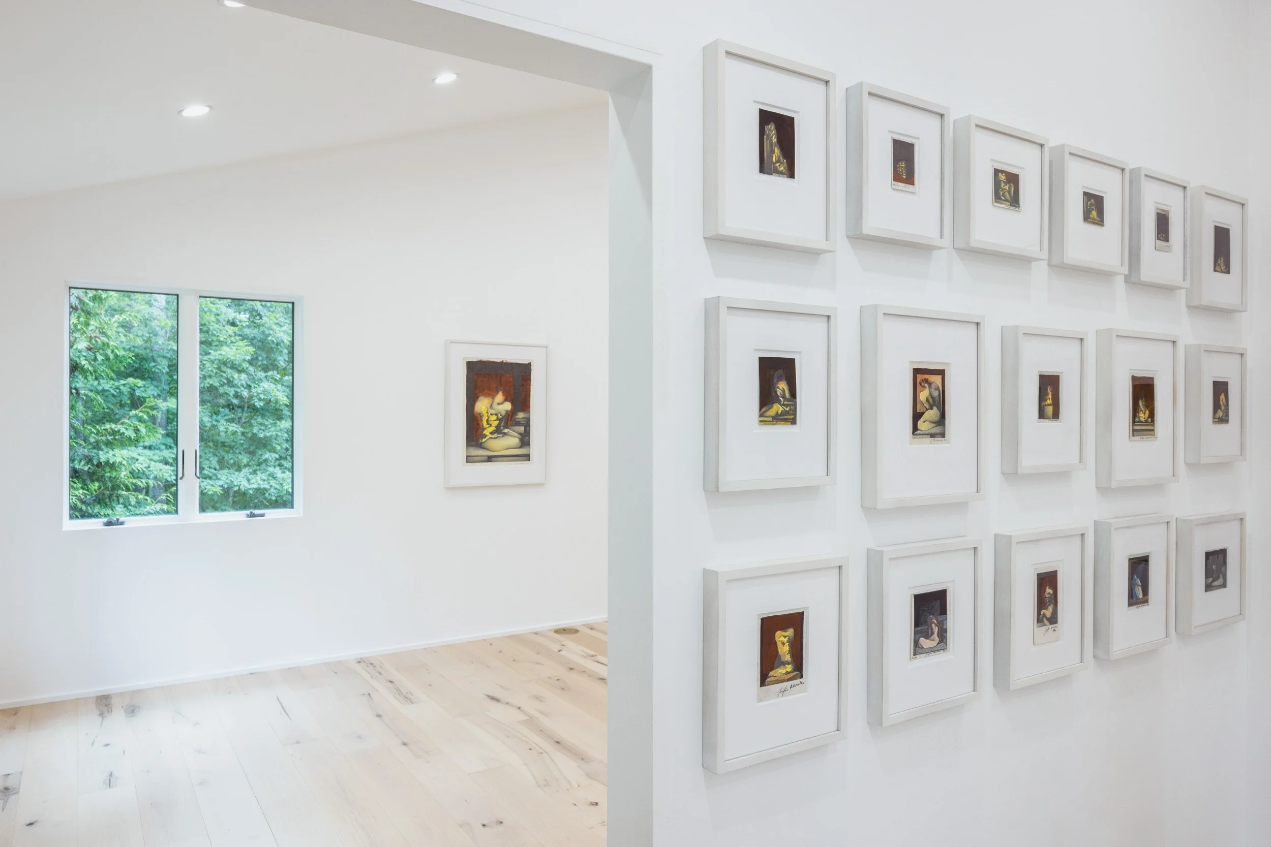 An art gallery displaying numerous framed artworks on a white wall. There are two large windows showing green foliage outside. The gallery has a light wood floor and bright lighting.