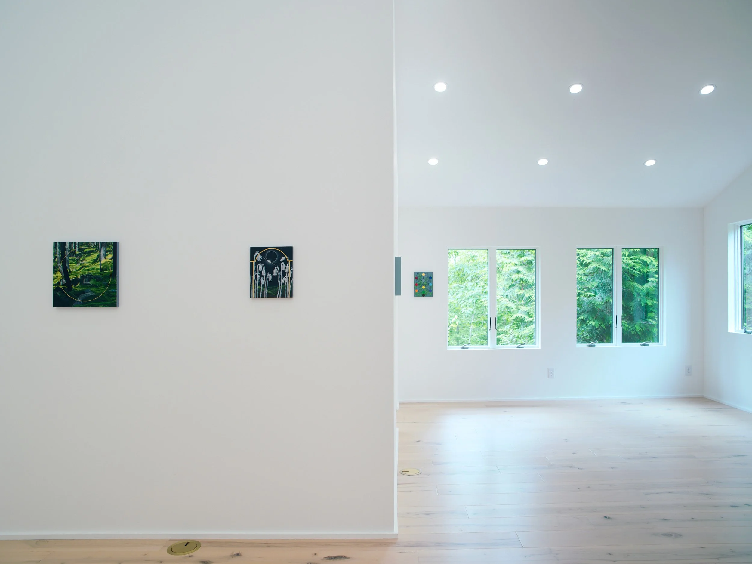 A modern, well-lit living space with large windows showing green trees outside, featuring white walls, light wooden flooring, and minimalistic artwork.