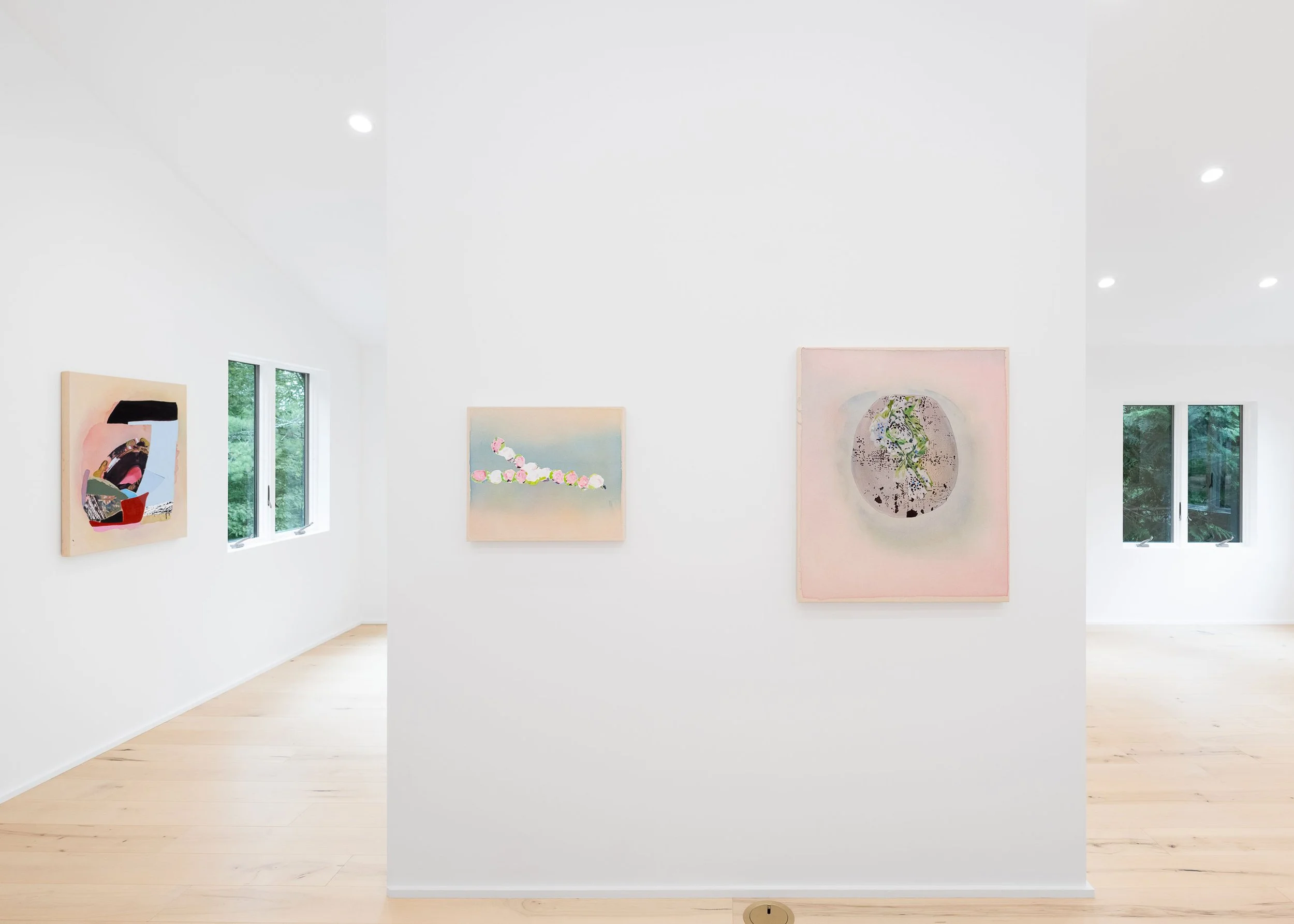 Contemporary art gallery with white walls and light wood flooring, featuring three framed artworks of abstract human figures hanging on a central white partition, with large windows showing greenery outside and a gray sofa on the right side.