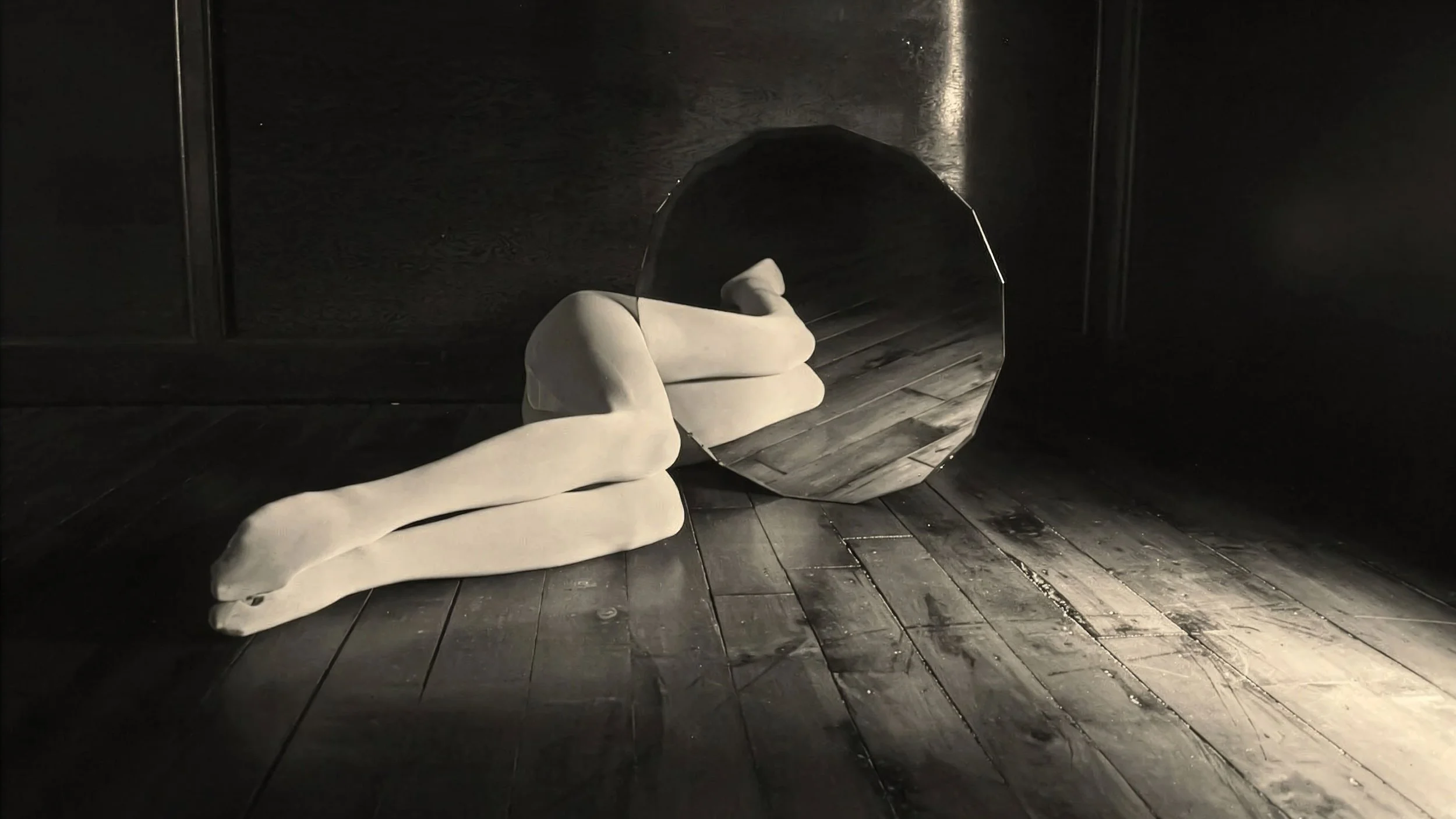A white fabric sculpture of a reclining figure lies on a dark wooden floor in front of a circular mirror reflecting part of the sculpture and the dark wall behind it.