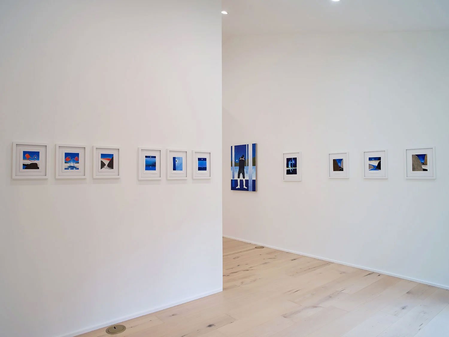 Art gallery with multiple framed abstract artworks on white walls, featuring blue, black, and red colors, and a central larger painting with a figure on a striped blue background.