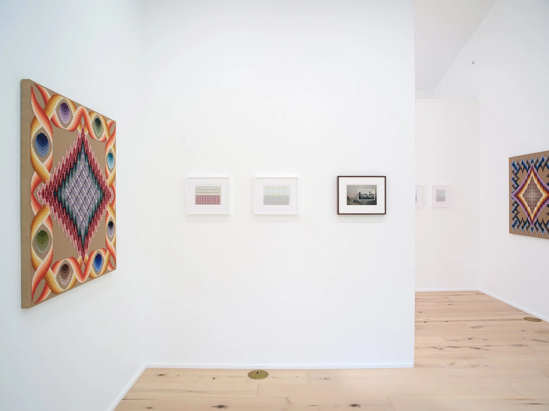 Art gallery with colorful textile artwork on the left and smaller framed photographs on the white wall, with a wooden floor and corner view of the room.