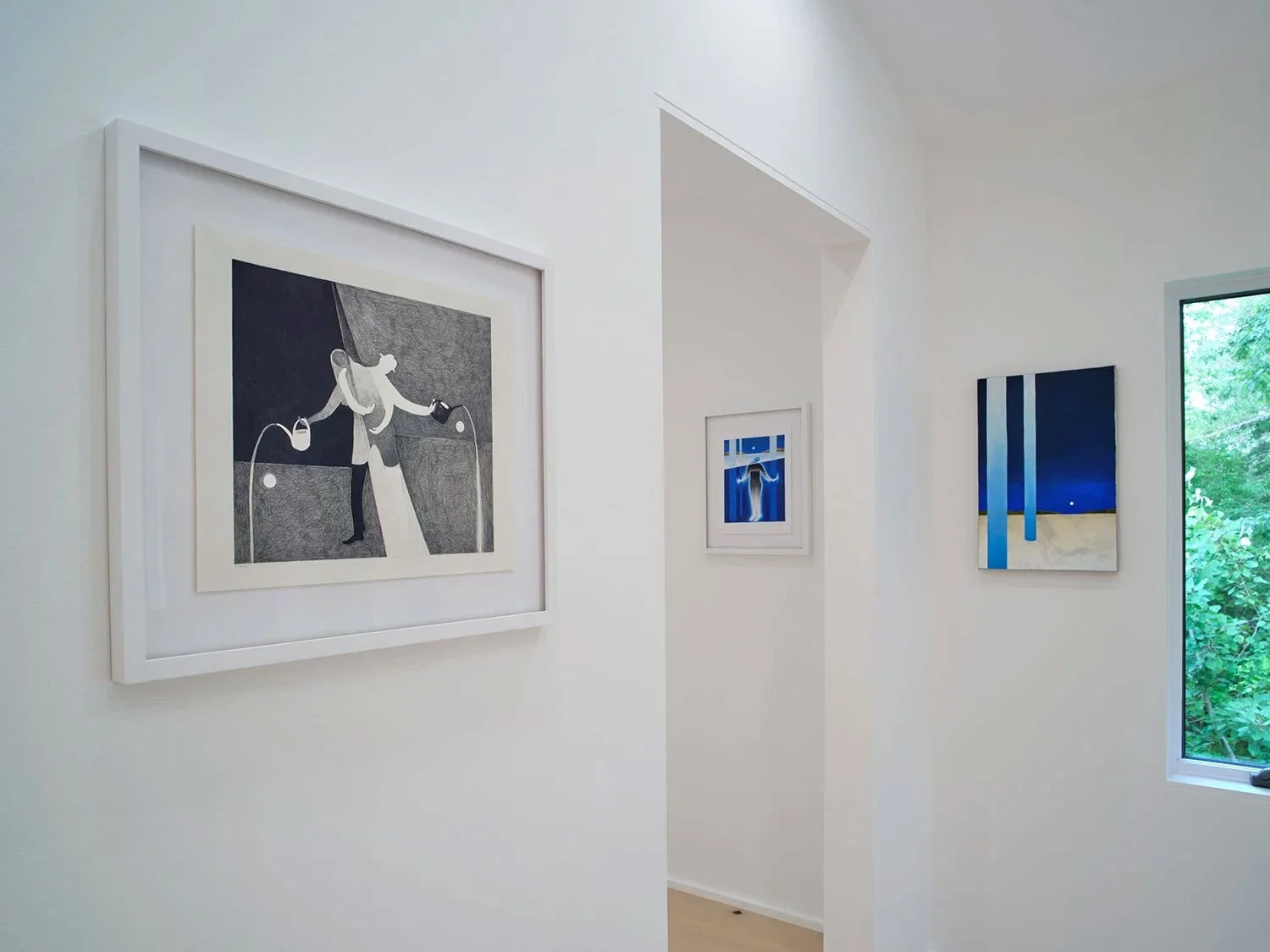White gallery wall with three framed artworks, a large window showing trees outside, and soft natural light