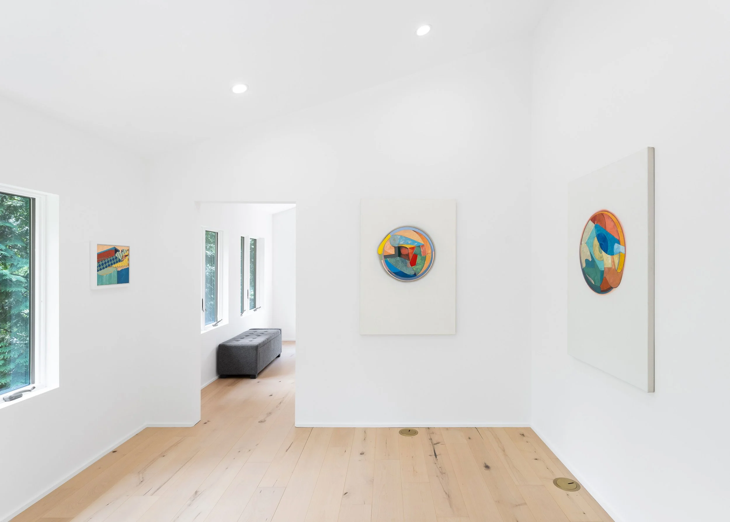 A minimalistic art gallery with white walls featuring abstract colorful paintings, windows with greenery outside, and light wood flooring.