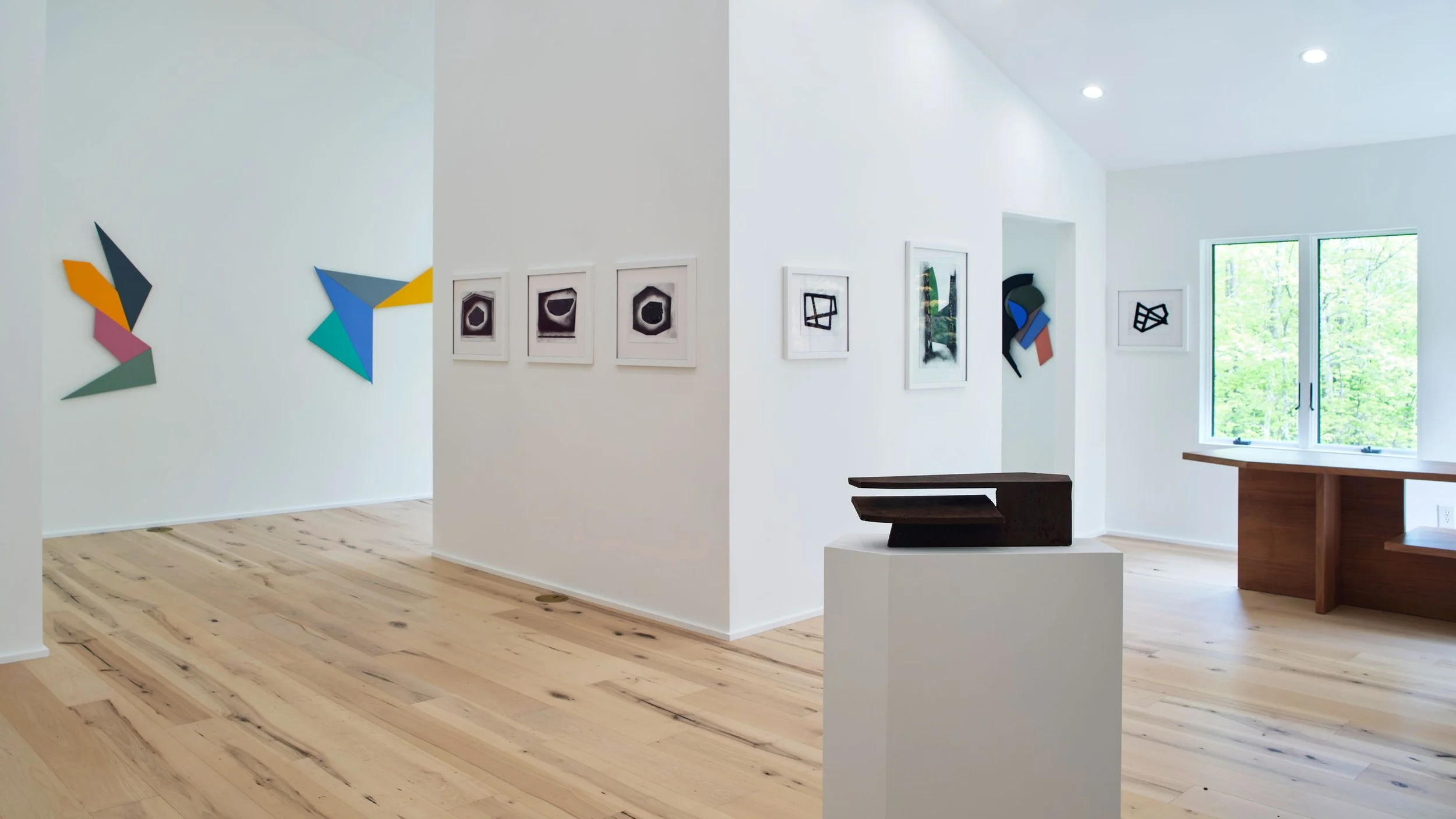 An art gallery with white walls and light wood flooring, displaying abstract artwork and framed black-and-white pieces, near a window overlooking greenery.
