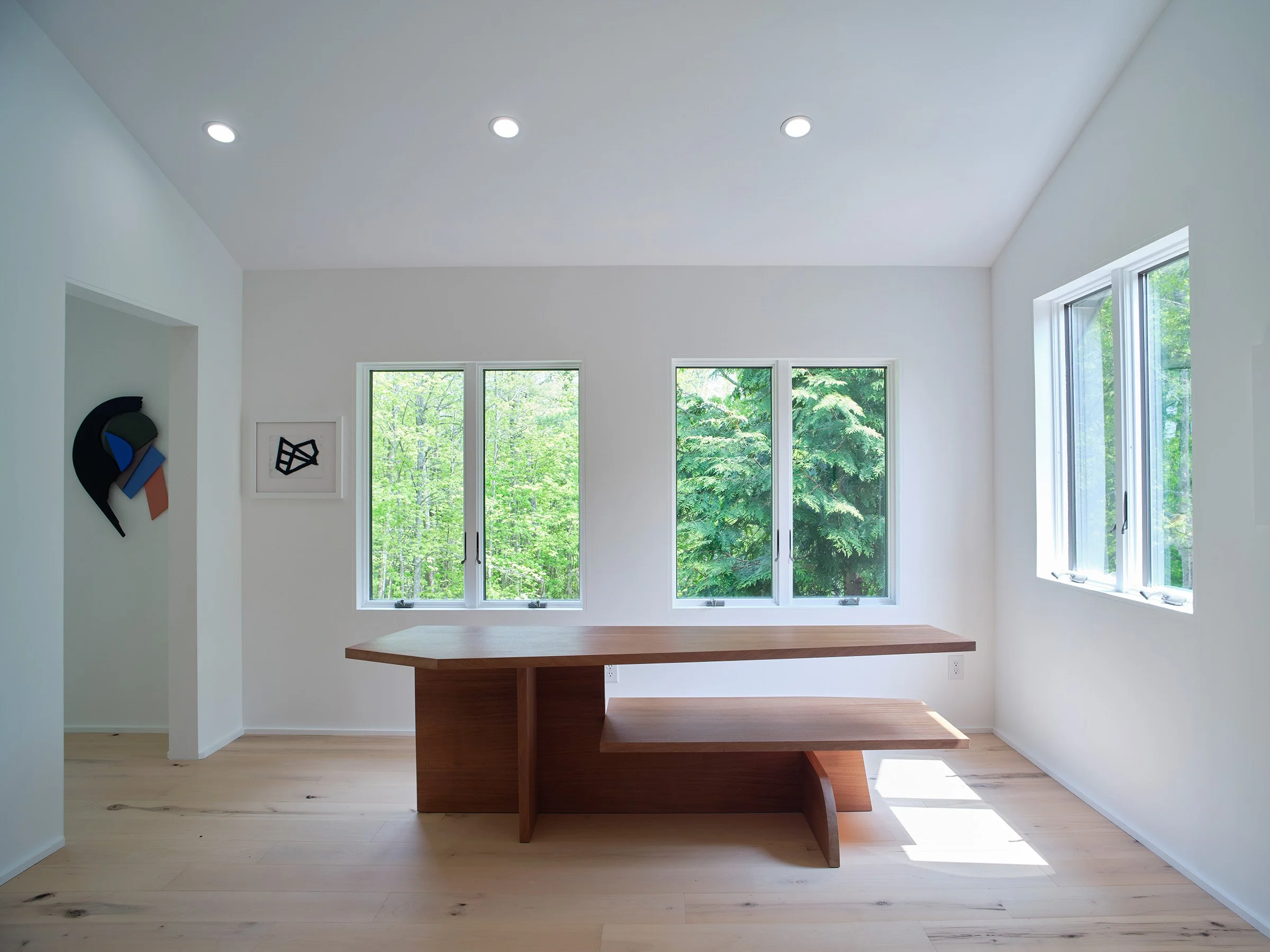 Minimalist room with large windows showing green trees outside, a wooden table with a bench, and abstract art on the wall.