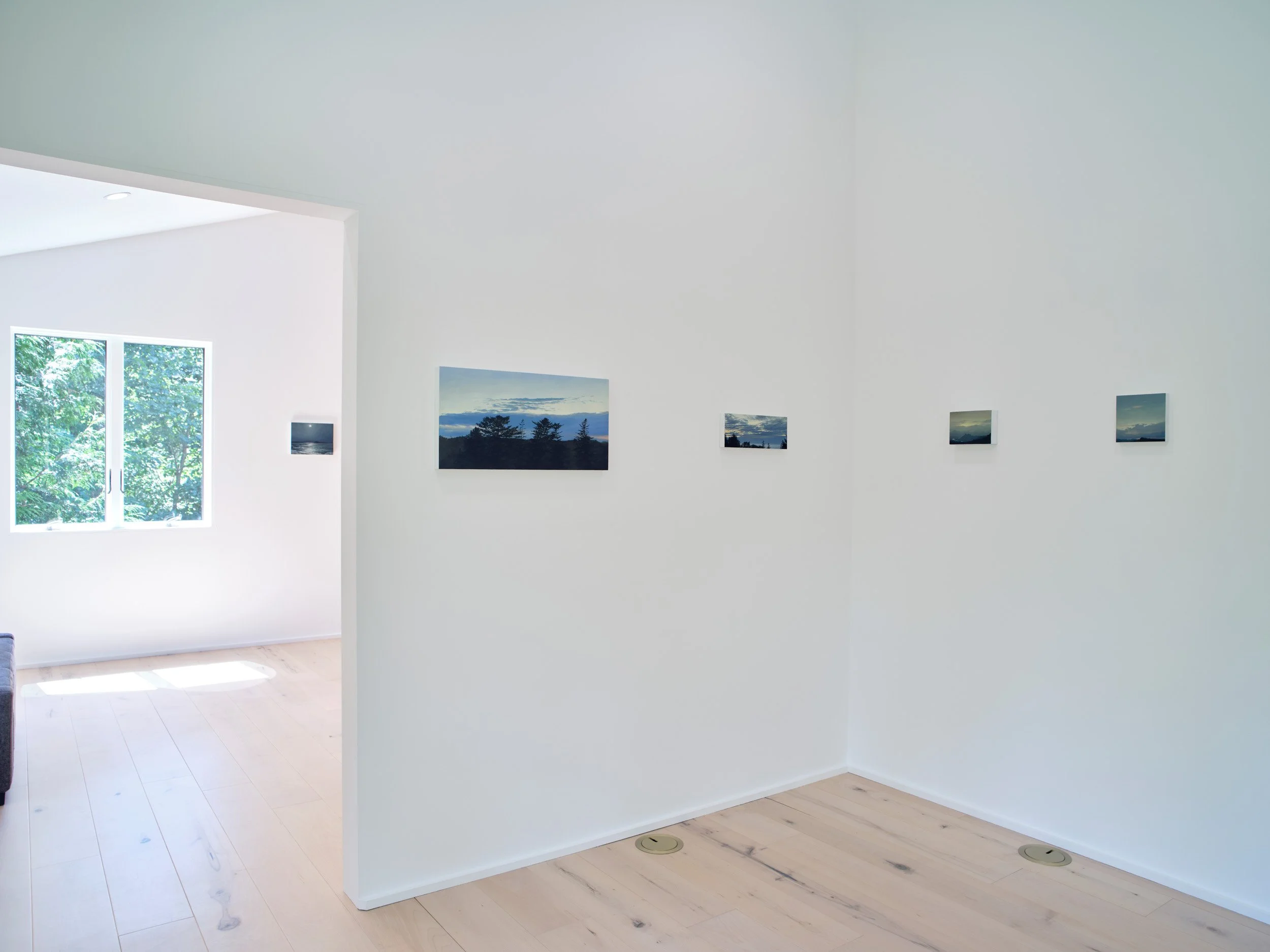 A white interior room with minimal decor, featuring a large window with greenery outside, and four landscape photographs hanging on the wall. The floor is light wood with visible knots and grain, and there are two circular floor vents.
