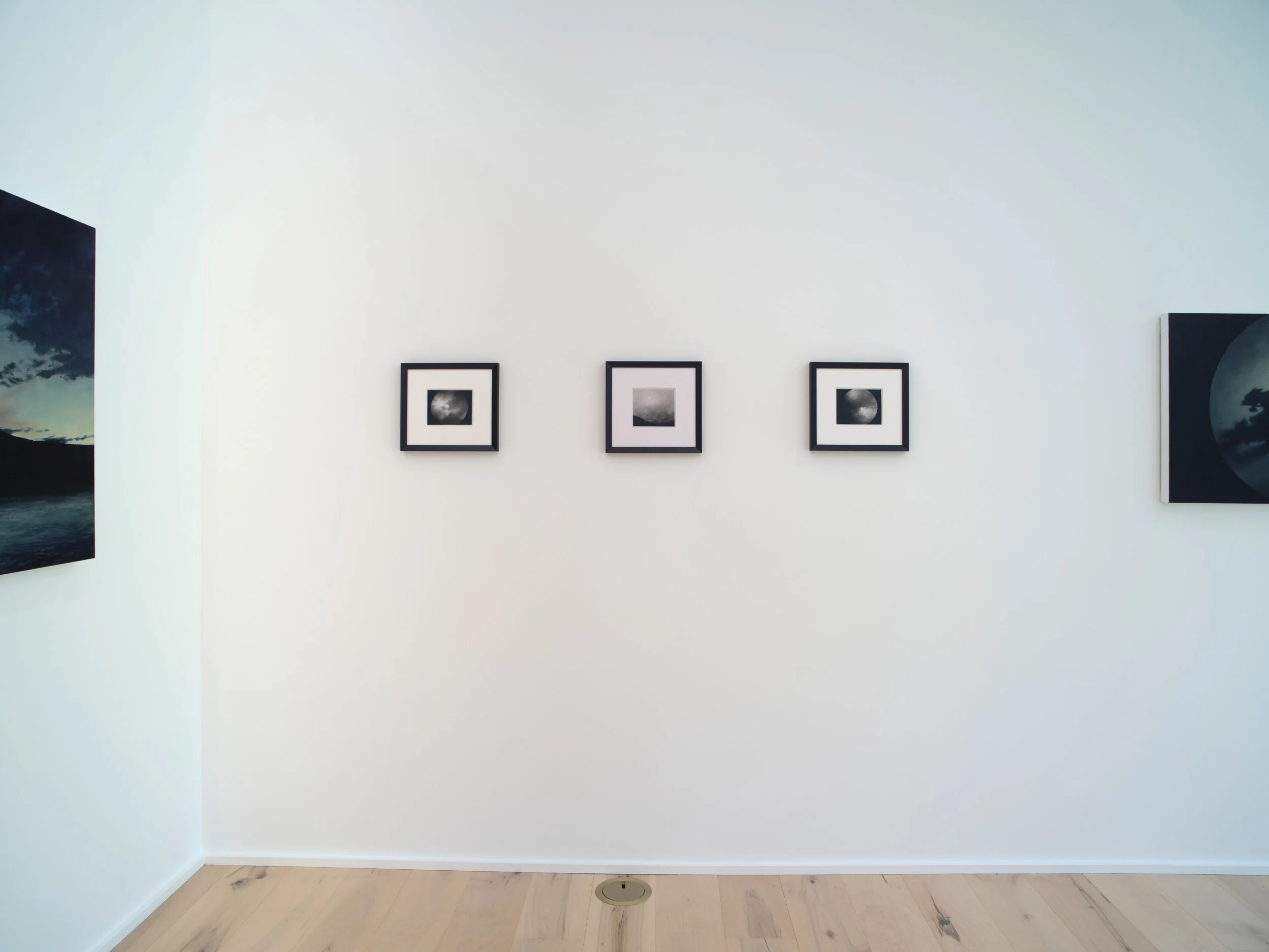 Empty white gallery wall with three small black and white framed photographs of the moon, and two large dark landscape photographs on either side, wooden floor with floor vent in the center.