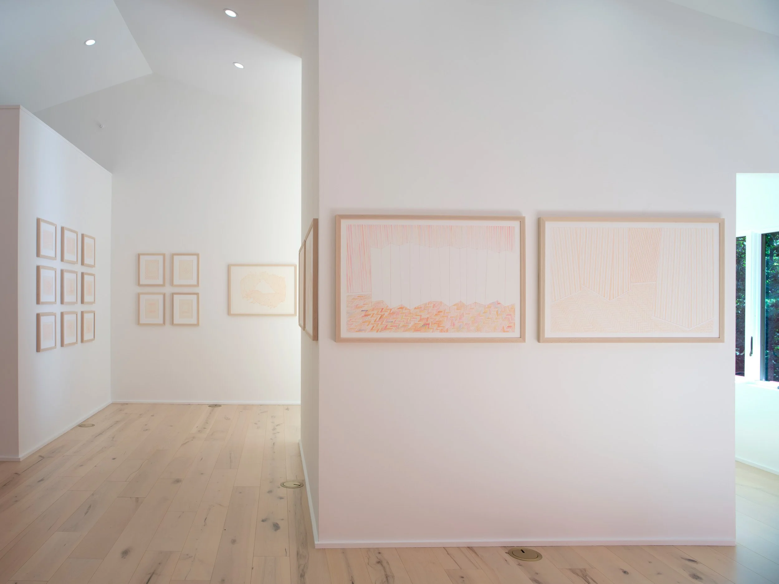 An interior view of an art gallery with white walls and light wooden flooring displaying framed artwork, with a door or window at the far end showing greenery outside.