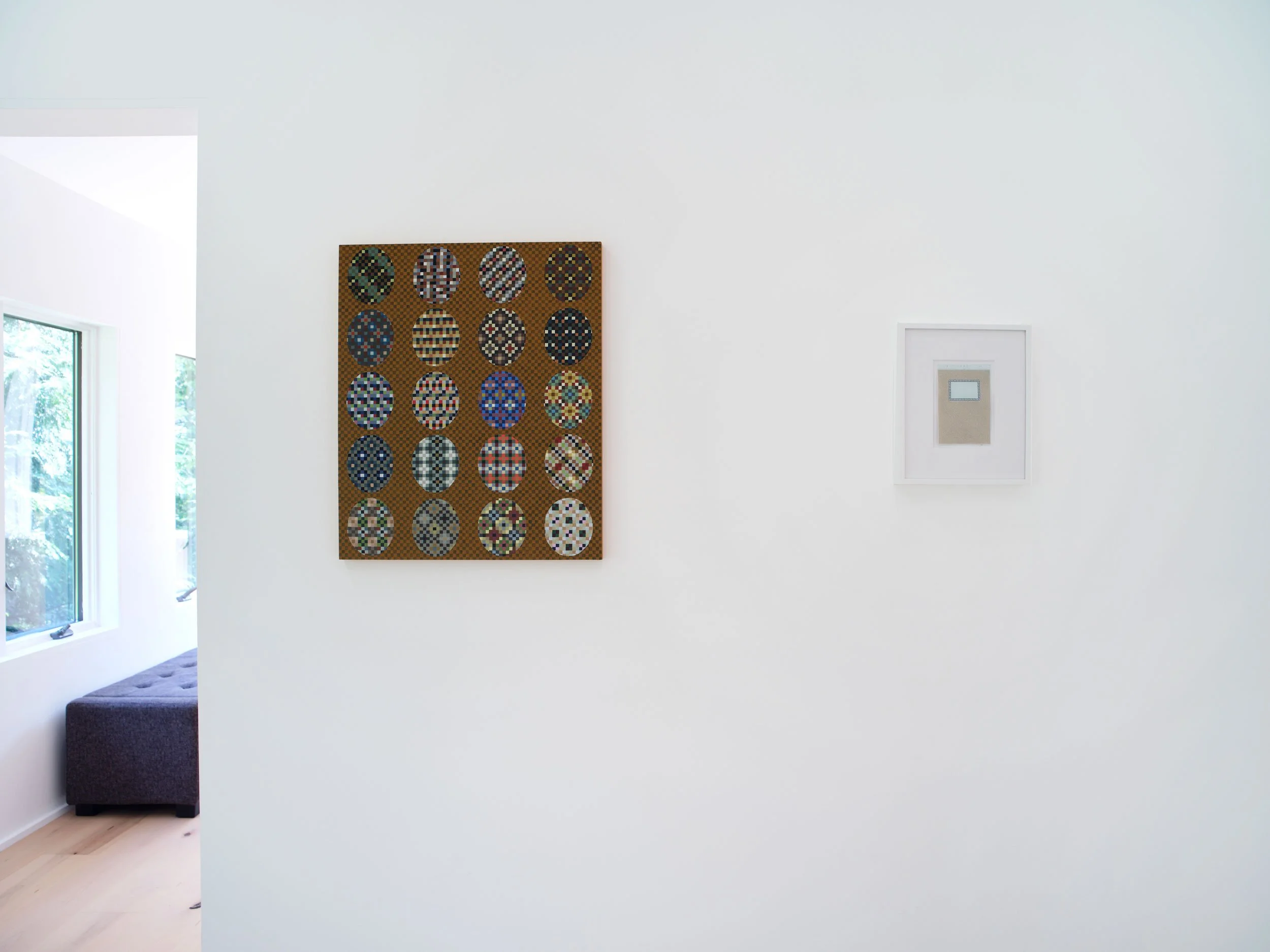 White gallery wall with a grid of brightly colored oval-patterned artworks and a small framed piece on the right side, next to a window with trees outside