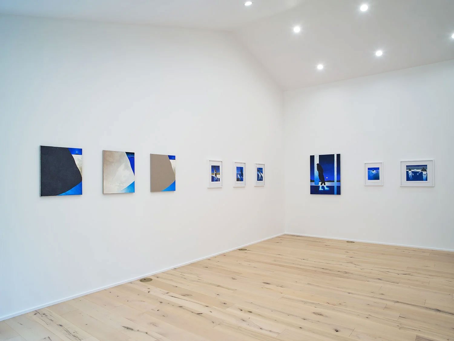 Art gallery with white walls displaying several framed and unframed photographs or paintings with blue tones, illuminated by ceiling lights, and a light wooden floor.
