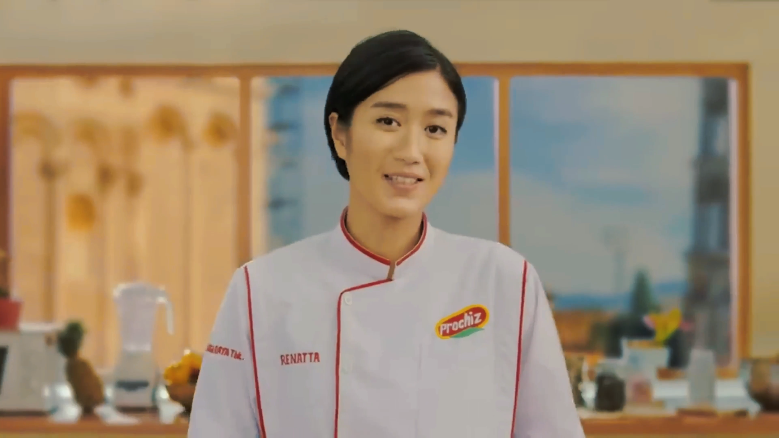 A woman in a white chef jacket with red trim and a Prochiz logo stands in a kitchen, smiling.