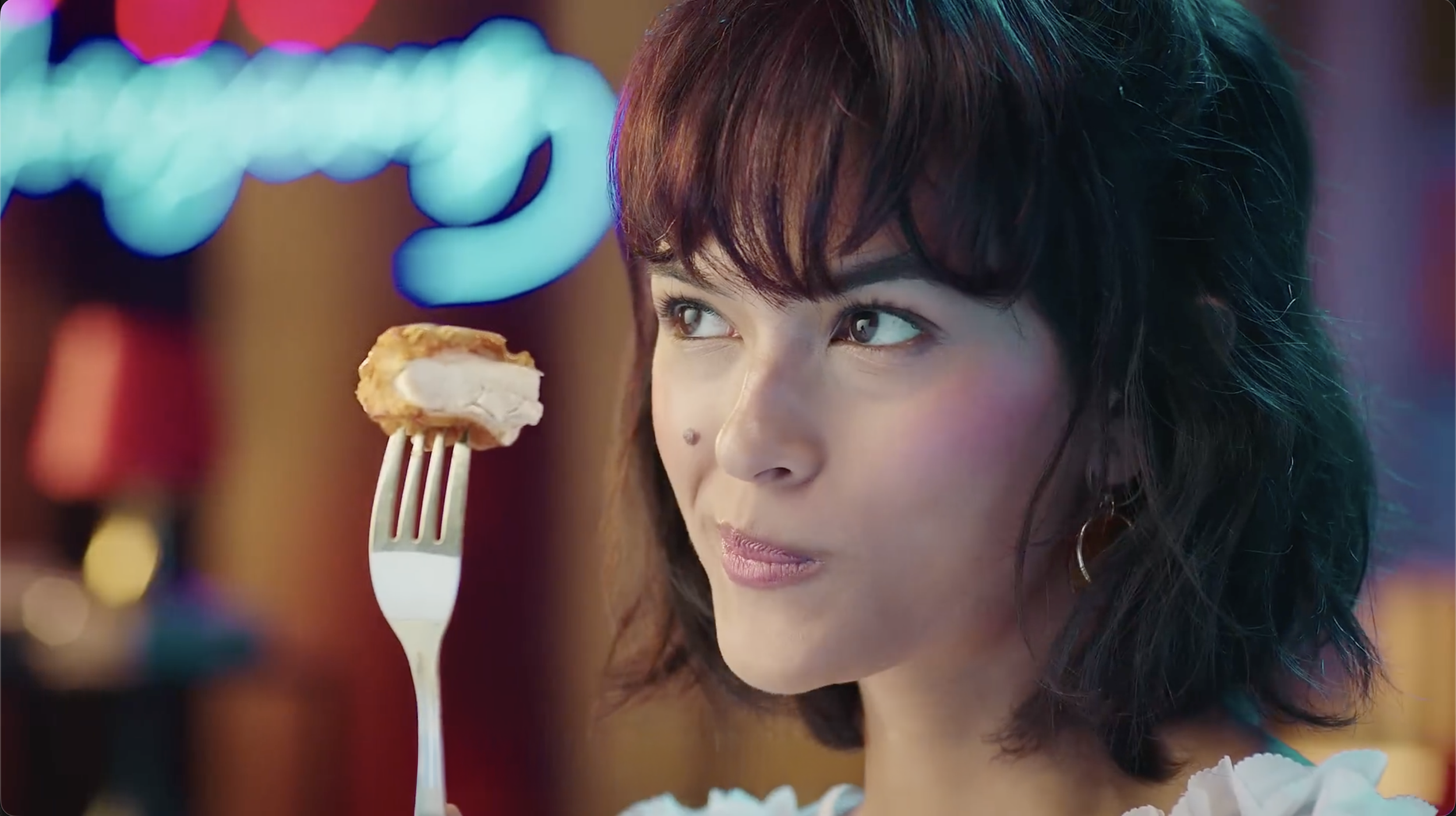 A woman with dark hair and a beauty mark on her cheek holds a fork with a piece of chicken near her mouth, in a setting with blurred colorful lights in the background.