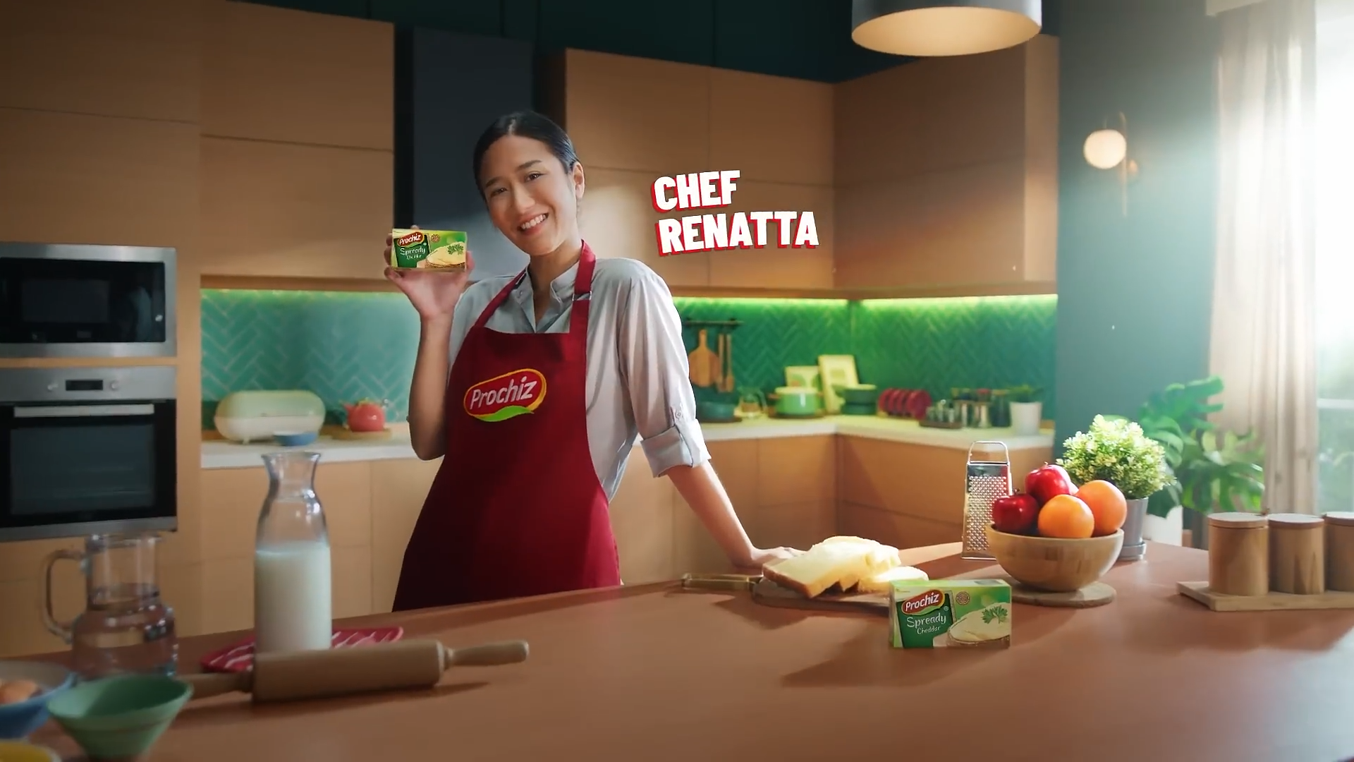 Woman in a kitchen holding a package of Prochiz spreadable cheddar cheese, smiling at the camera, with fresh fruits and kitchen utensils on the counter.