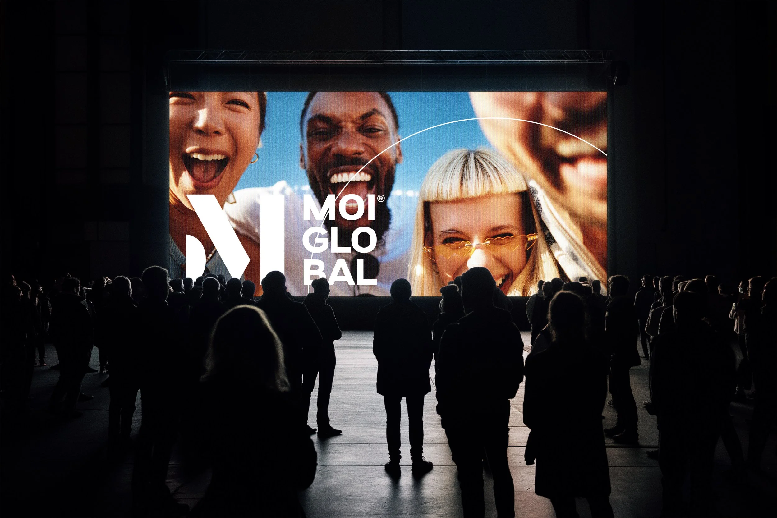 Silhouettes of people watching a large outdoor screen displaying a close-up of smiling faces in bright daylight.