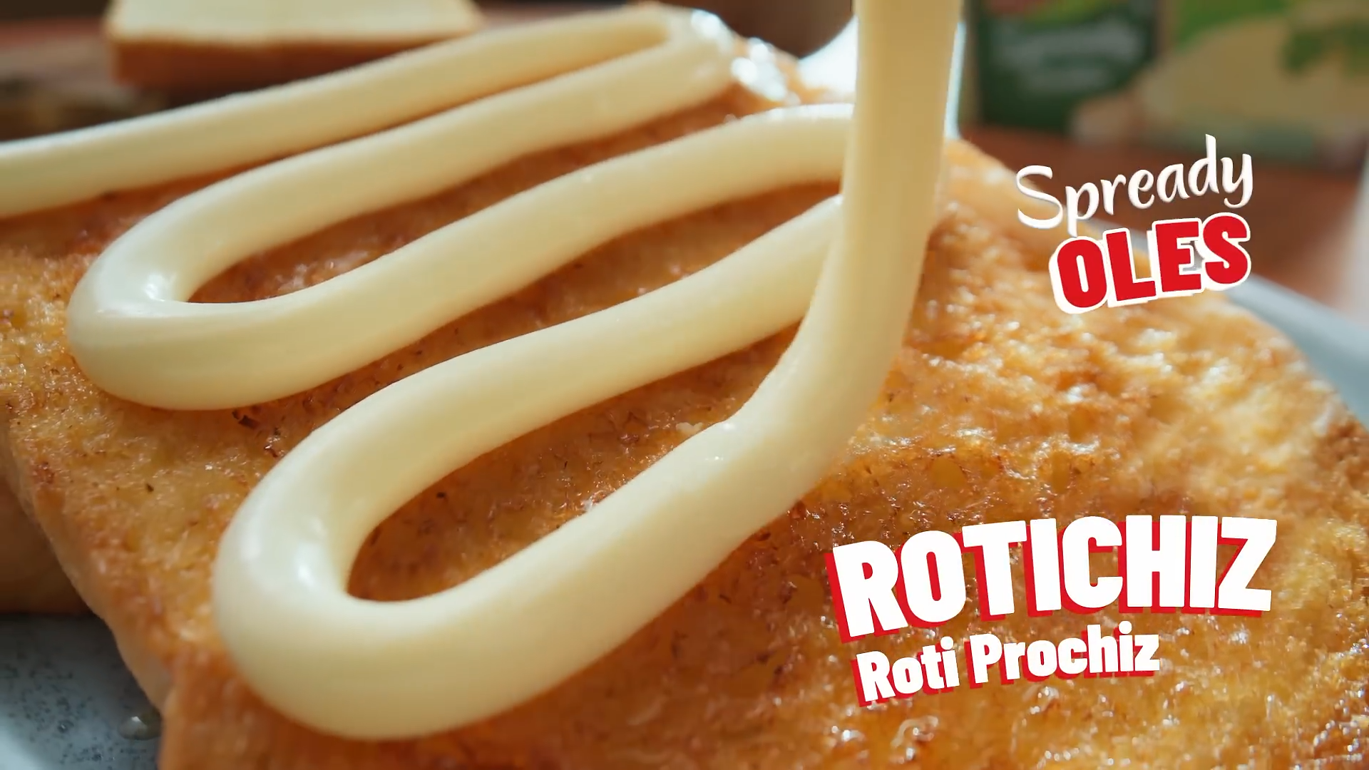 Close-up of a breaded fried food item with white mayonnaise zigzagged on top, with text overlay that reads 'Spready OLES Roti Prochiz'.