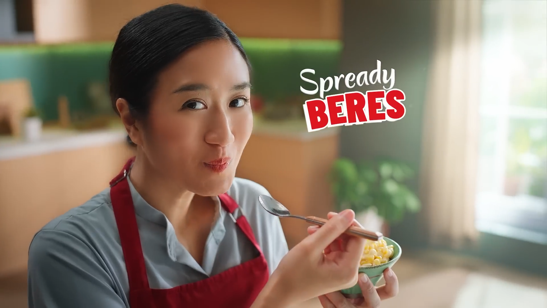 A woman with dark hair in a bun wearing a gray shirt and red apron, holding a spoon and a bowl of creamy corn, smiling in a cozy kitchen with sunlight and green plants.