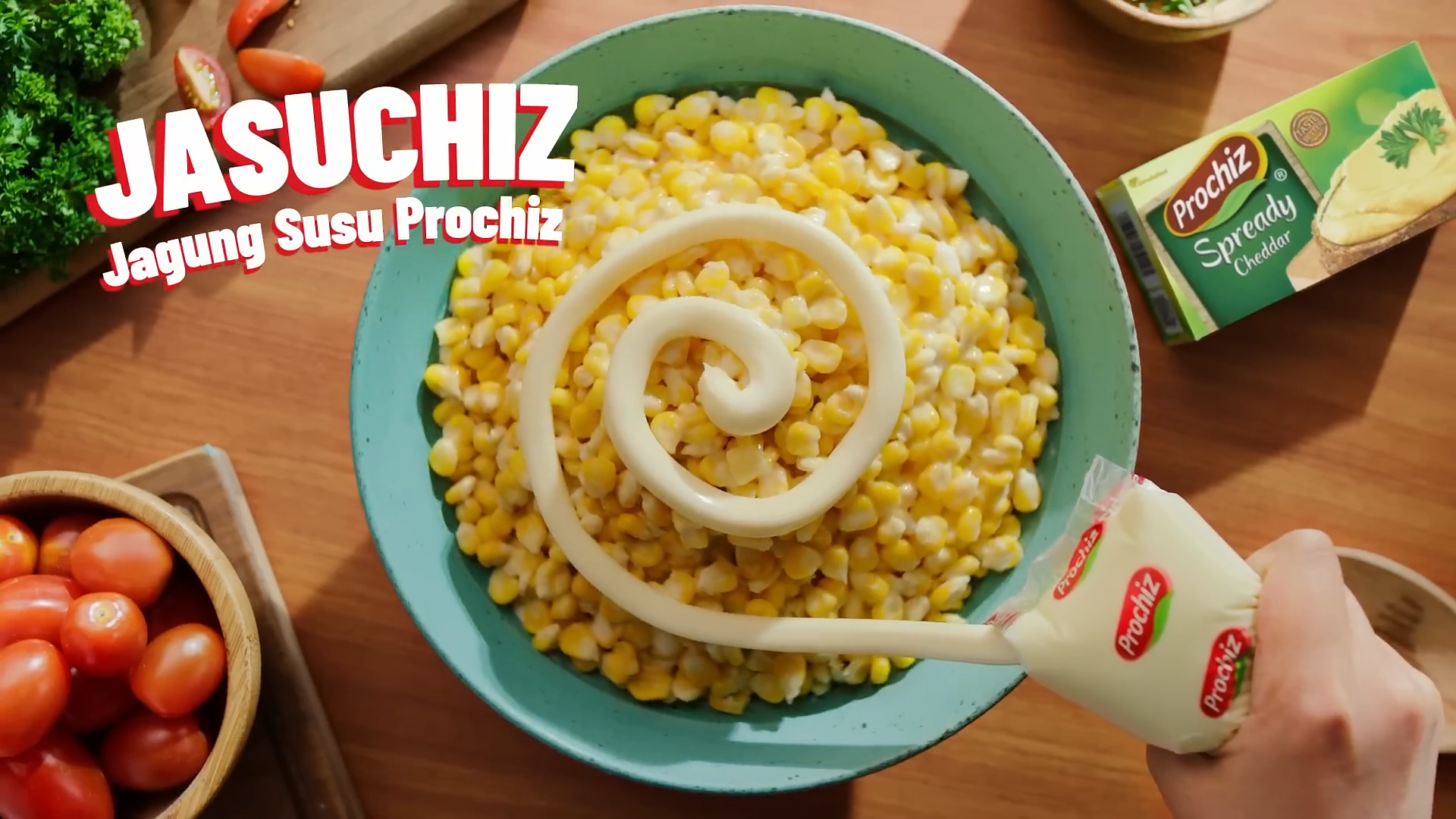 A bowl of sweet corn salad garnished with a swirl of mayonnaise. A hand is squeezing Prochiz spread cheese onto the salad. Surrounding the bowl are tomatoes, fresh herbs, and a box of Prochiz spread cheese on a wooden table.