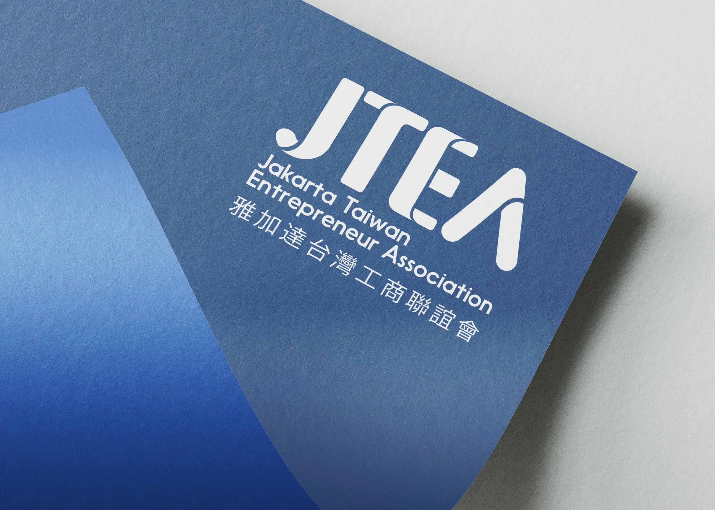 Close-up of a blue conference or association card with white text that reads "JTEA", "Jakarta Taiwanese Entrepreneur Association", and additional Chinese characters underneath, lying on a gray surface.