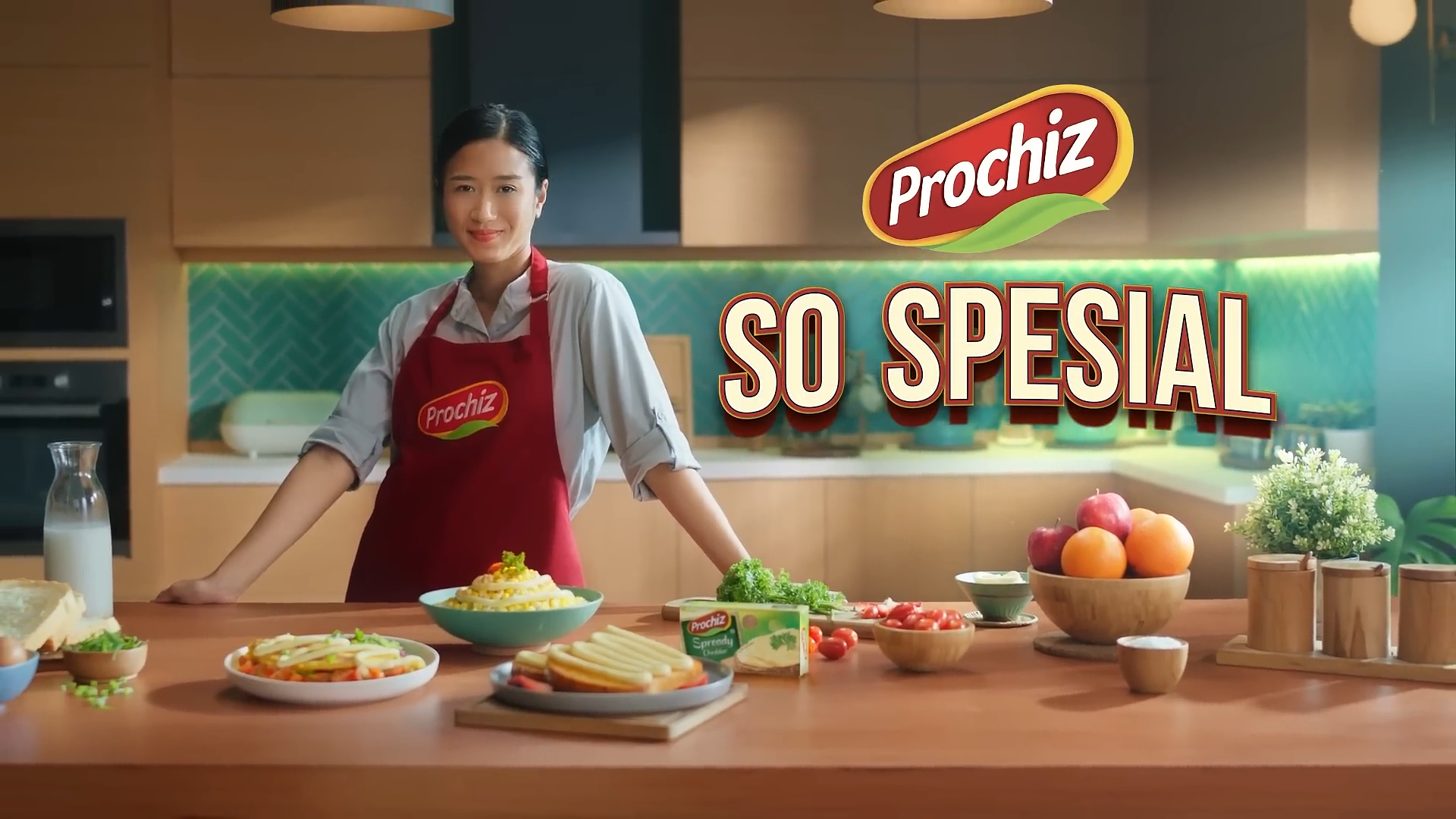 A woman wearing a red apron with the Prochiz logo, smiling in a kitchen with various foods on the table, with the words 'Prochiz SO SPESIAL' in bold text.