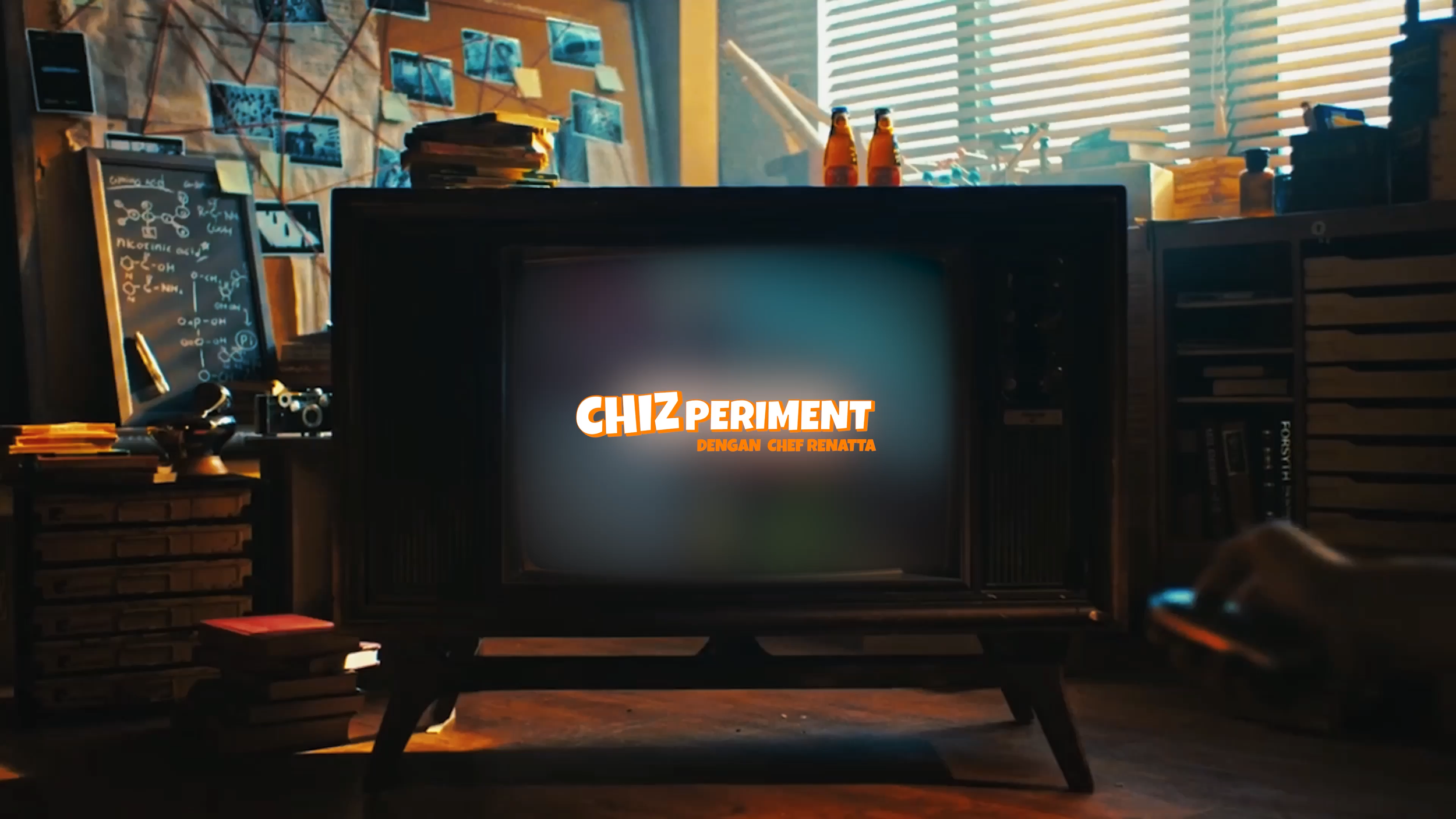 A cozy, cluttered workspace featuring a television with the text 'CHIZ PERIMENT DENGAN CHEF RENATTA' on the screen, surrounded by books, a blackboard with chemical equations, stacks of papers, and photographs on a wall, with sunlight streaming through blinds.