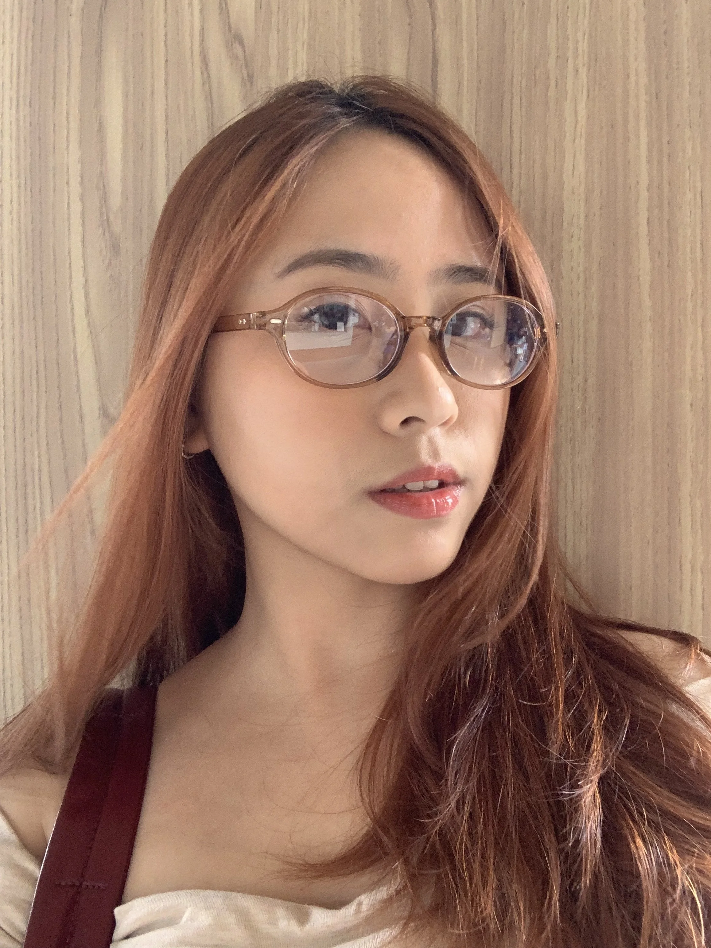 A young woman with long, reddish-brown hair, wearing round glasses with a transparent frame, looking slightly to the side, in front of a light-colored wooden background.