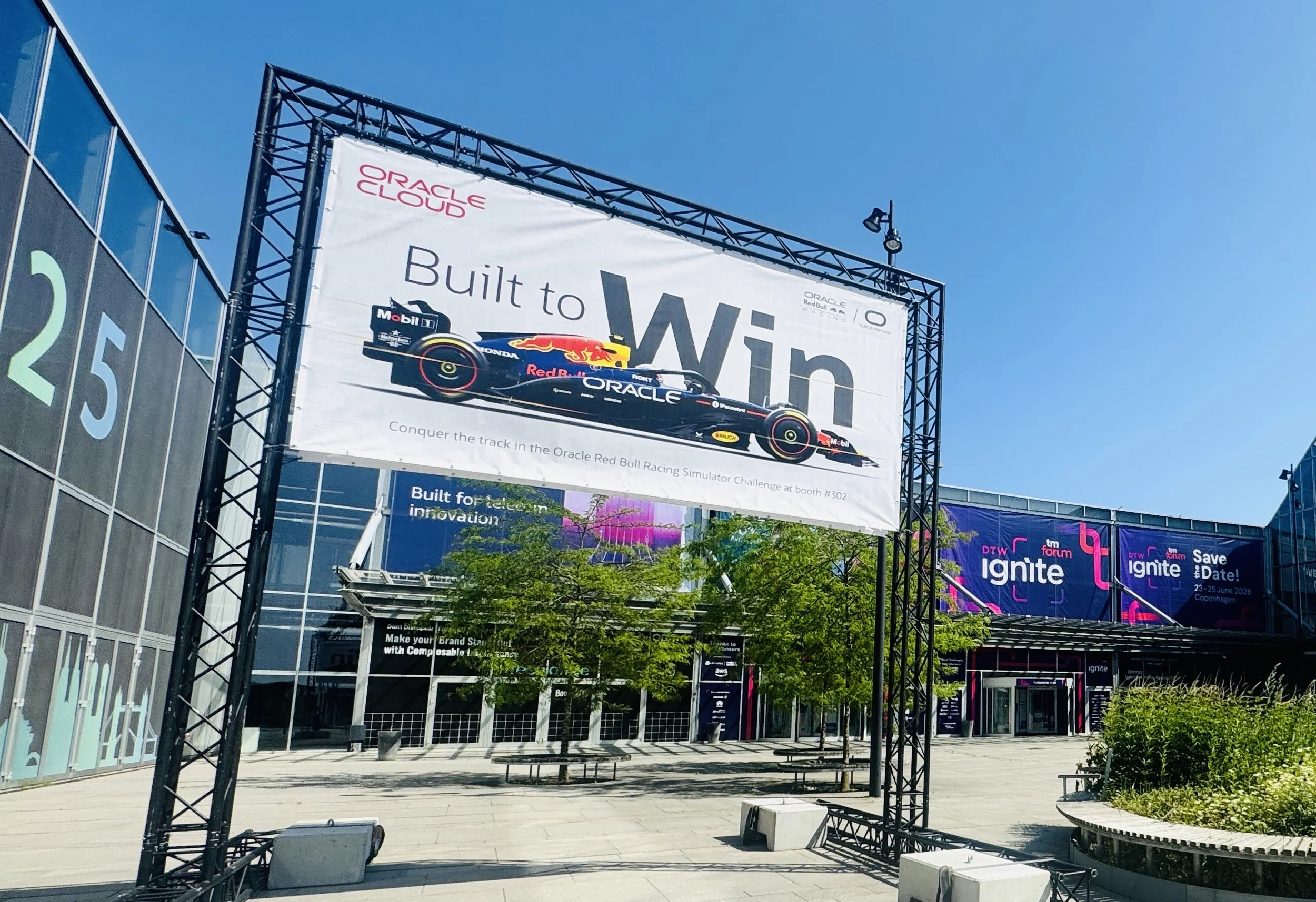 Billboard advertising Oracle Cloud services and Red Bull Racing Formula 1 car at an outdoor event, with a clear blue sky and modern buildings in the background.