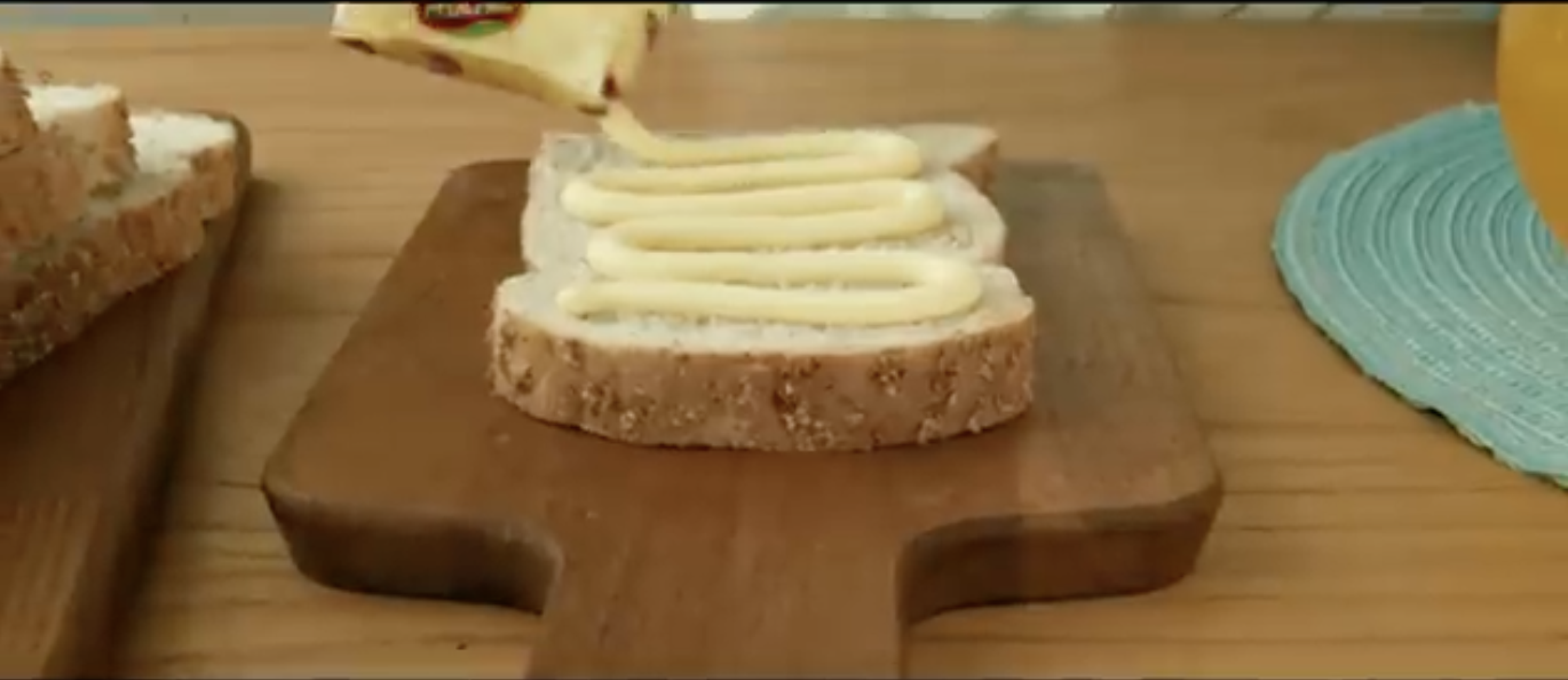 Slice of bread with mayonnaise being spread on it with a spreader on a wooden cutting board.