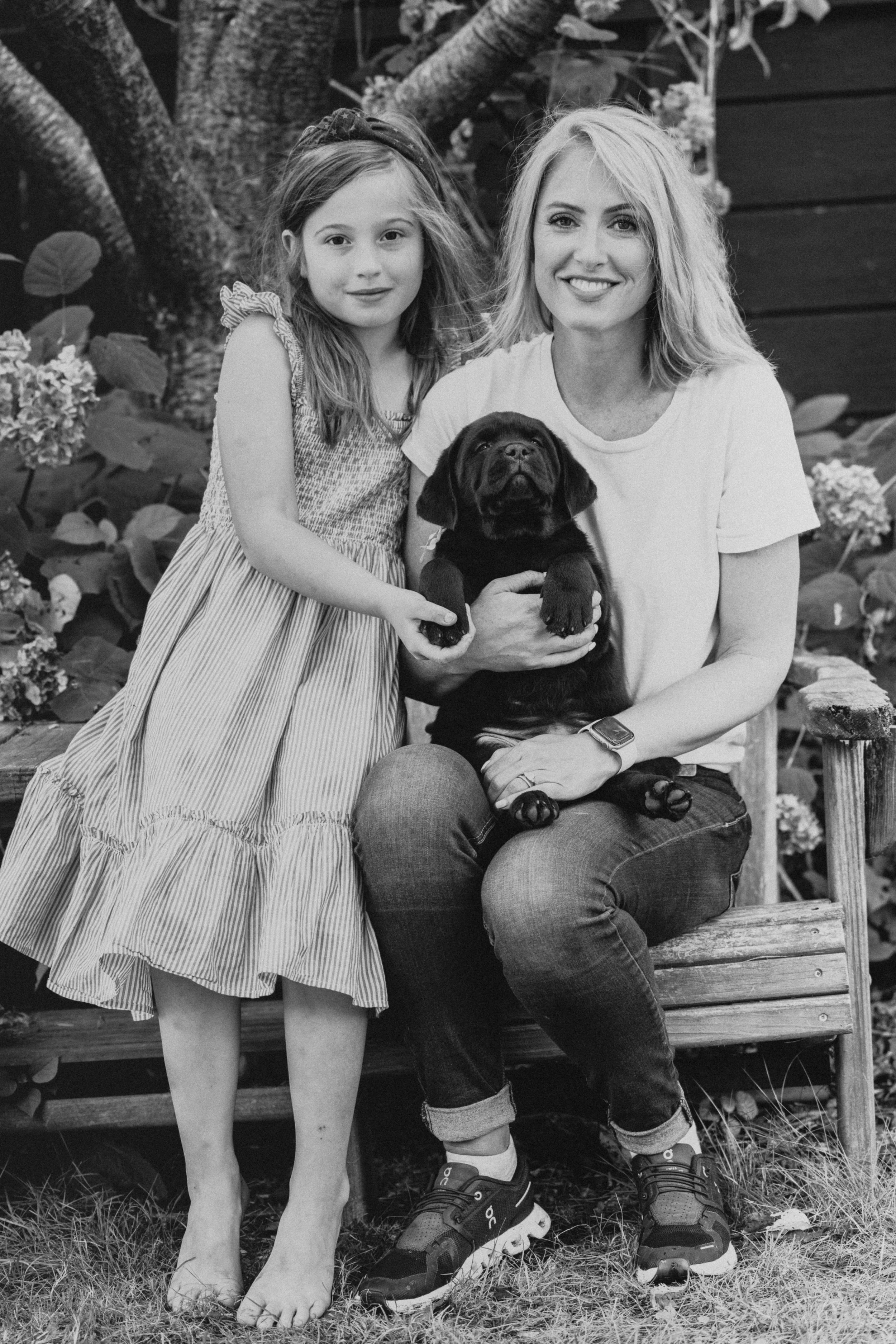 sitting outdoors on a wooden bench with a puppy. The woman is smiling, and the girl has a neutral expression. The woman is dressed casually in a t-shirt and jeans, while the girl is in a striped dress. The puppy is being held by the woman. The backgr